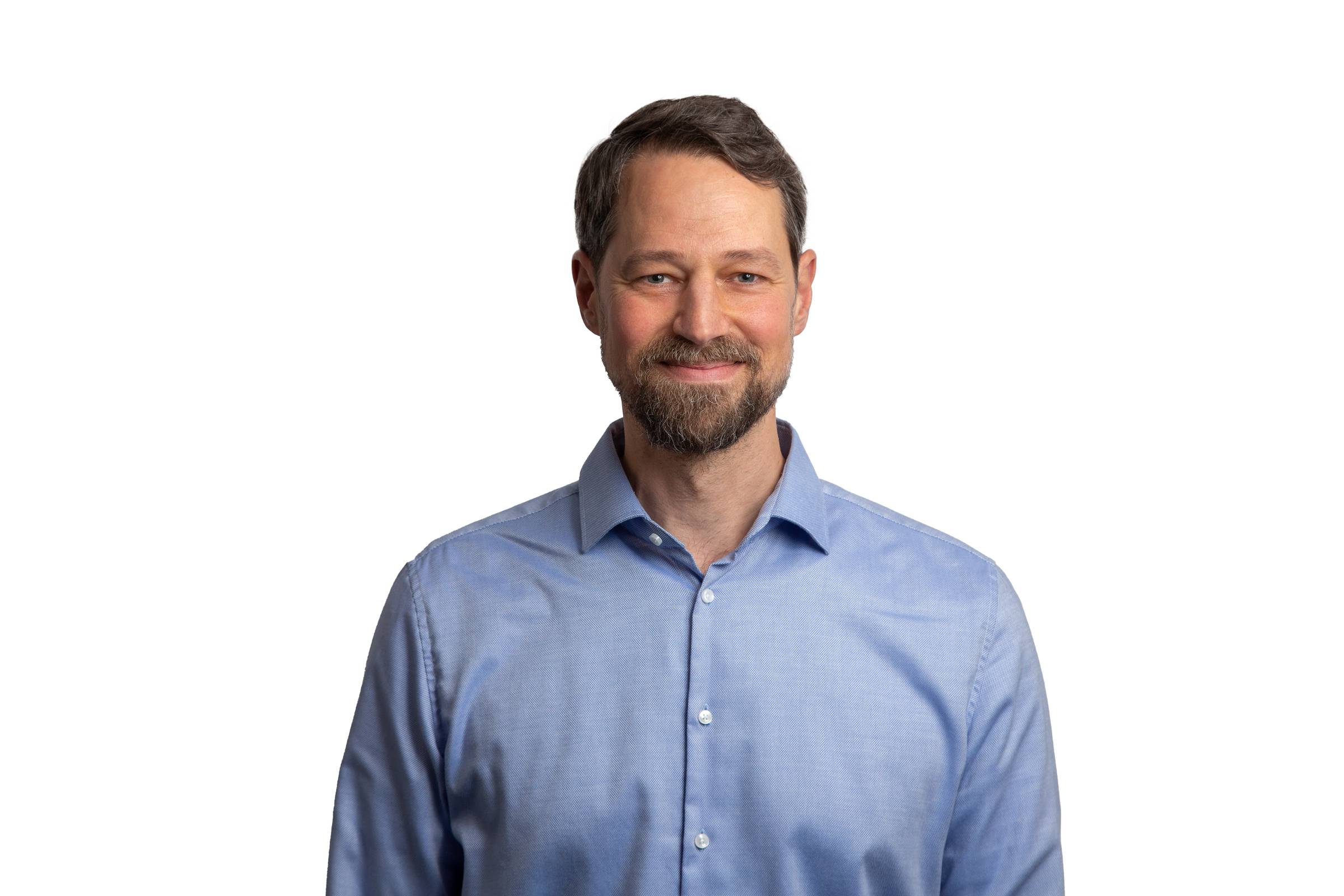 A man with a beard is smiling, wearing a light blue button-up shirt. The background is plain white.