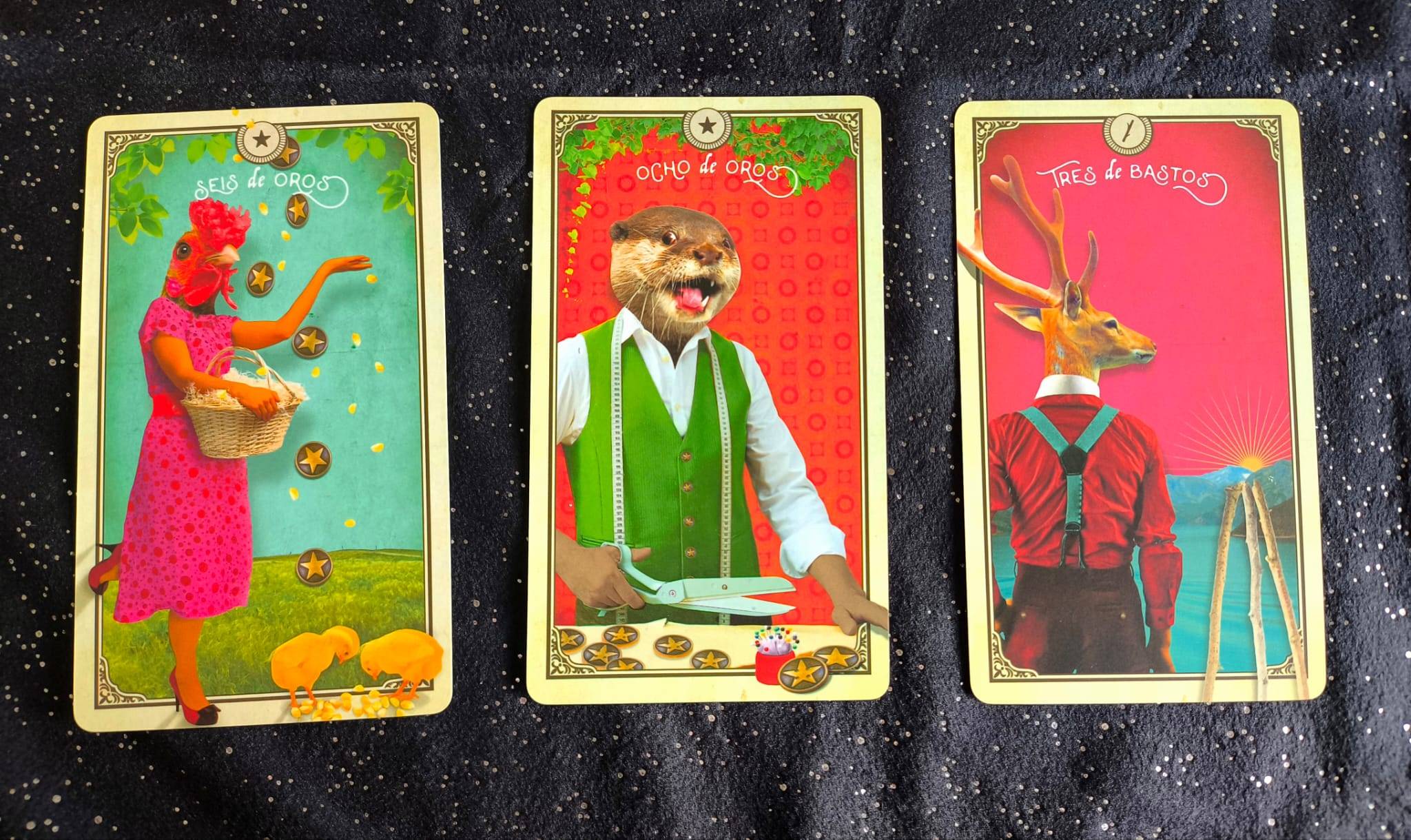 Sobre un tapete negro con pintas plateadas, hay tres cartas de tarot en las que las figuras representadas tienen cuerpo de persona y cabeza de animal, como un collage. El seis de oros tiene una mujer con cabeza de gallina dando de comer a unos pollitos, e