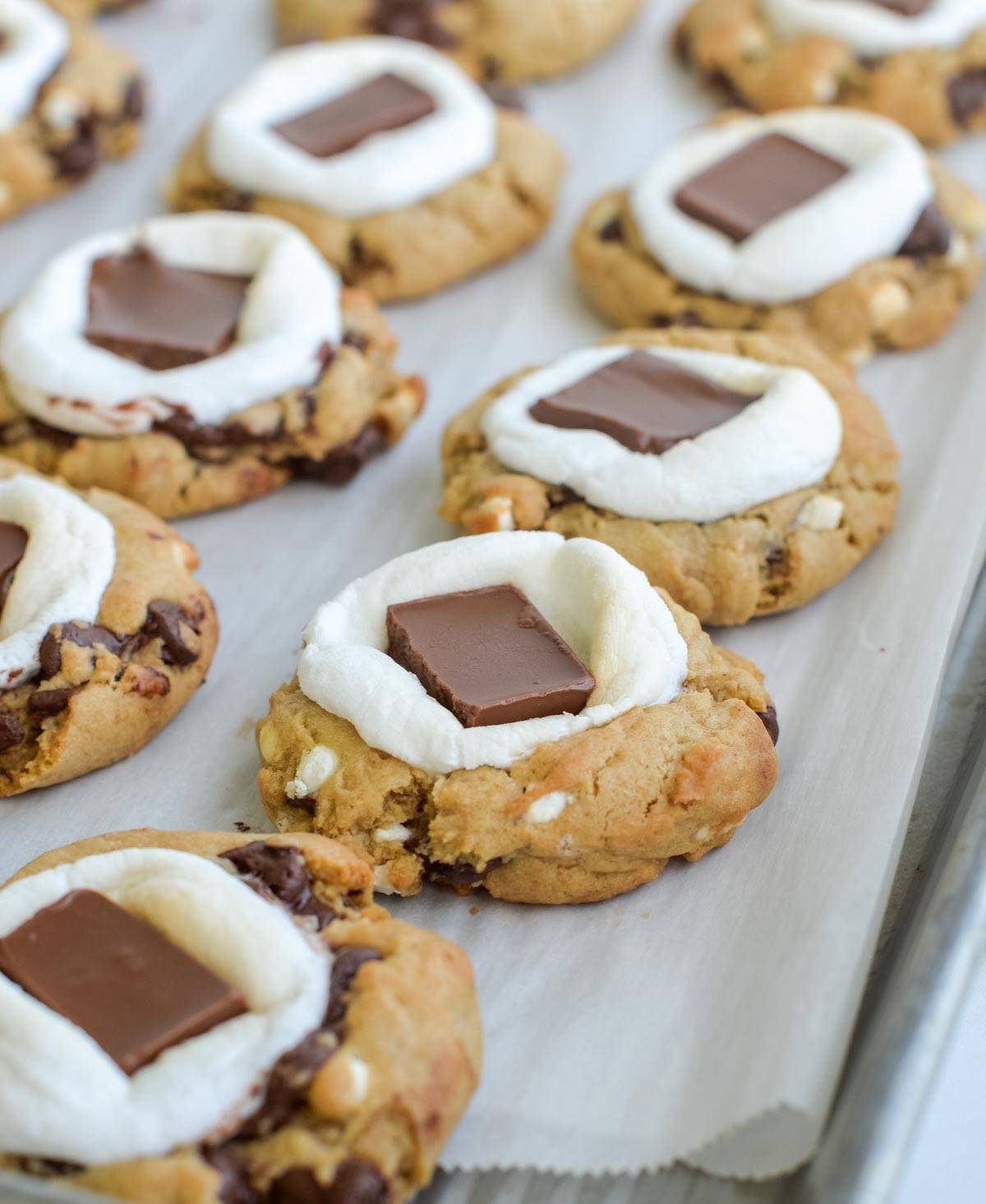 S'mores cookies topped with marshmallow and chocolate squares sit on a parchment lined cookie sheet.