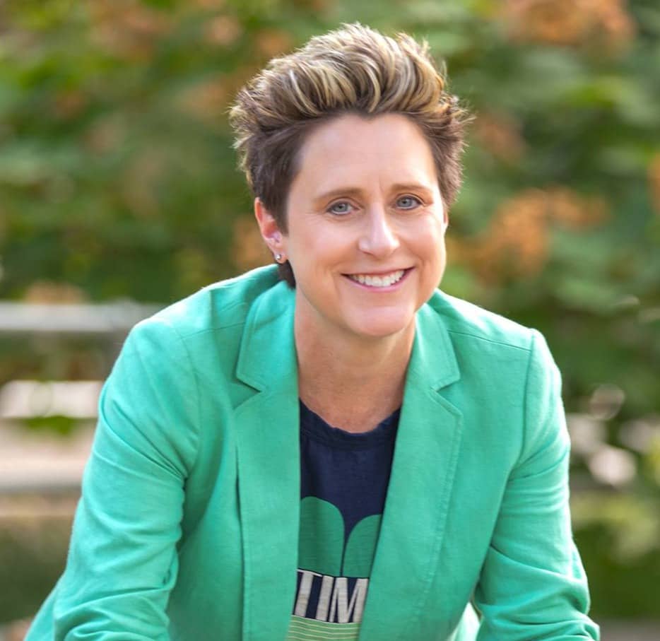 A person with short, styled hair is smiling at the camera. They are wearing a bright green blazer over a dark shirt. The background is blurred with natural greenery, suggesting an outdoor setting.