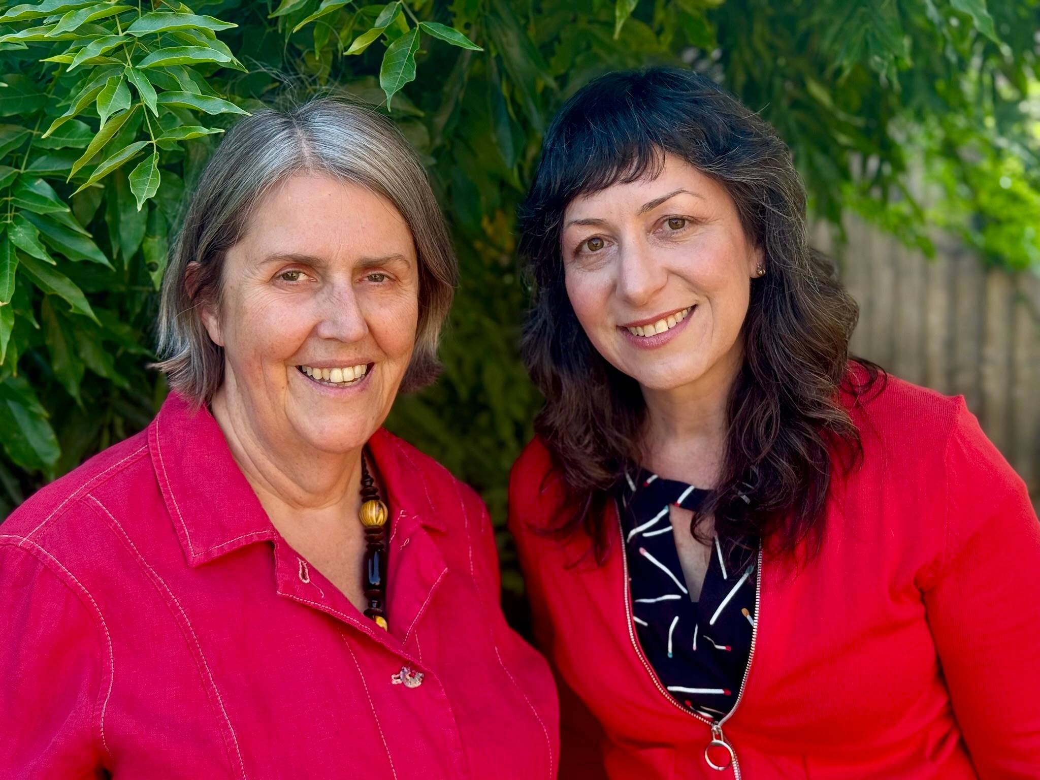 Claire and Kim are smiling at the camera, wearing red jackets. Green leaves and a wooden fence are in the background.