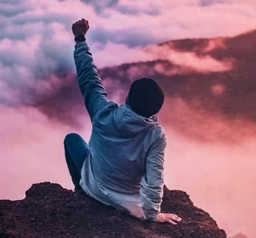 man sitting on mountain cliff facing white clouds rising one hand at golden hour