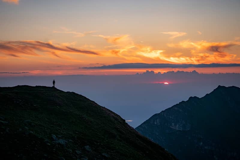 Person silhouetted on a mountain peak during a vibrant sunset.