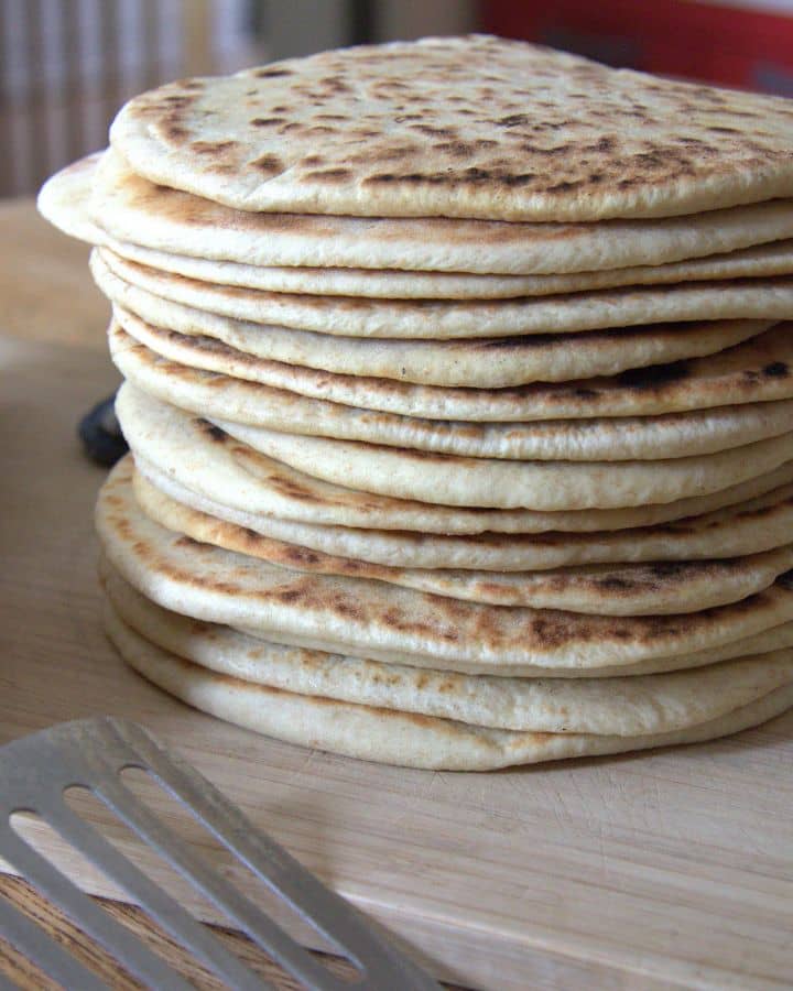 A tall stack of homemade flatbreads with golden brown spots, resting on a wooden cutting board. A metal spatula lies nearby.