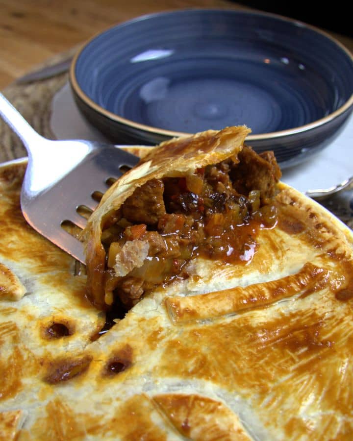 A close-up of a golden-brown beef pot pie with a flaky puff pastry crust. A fork lifts a slice, revealing a savory filling of beef, onions, and vegetables in a rich gravy. A blue bowl is in the background.