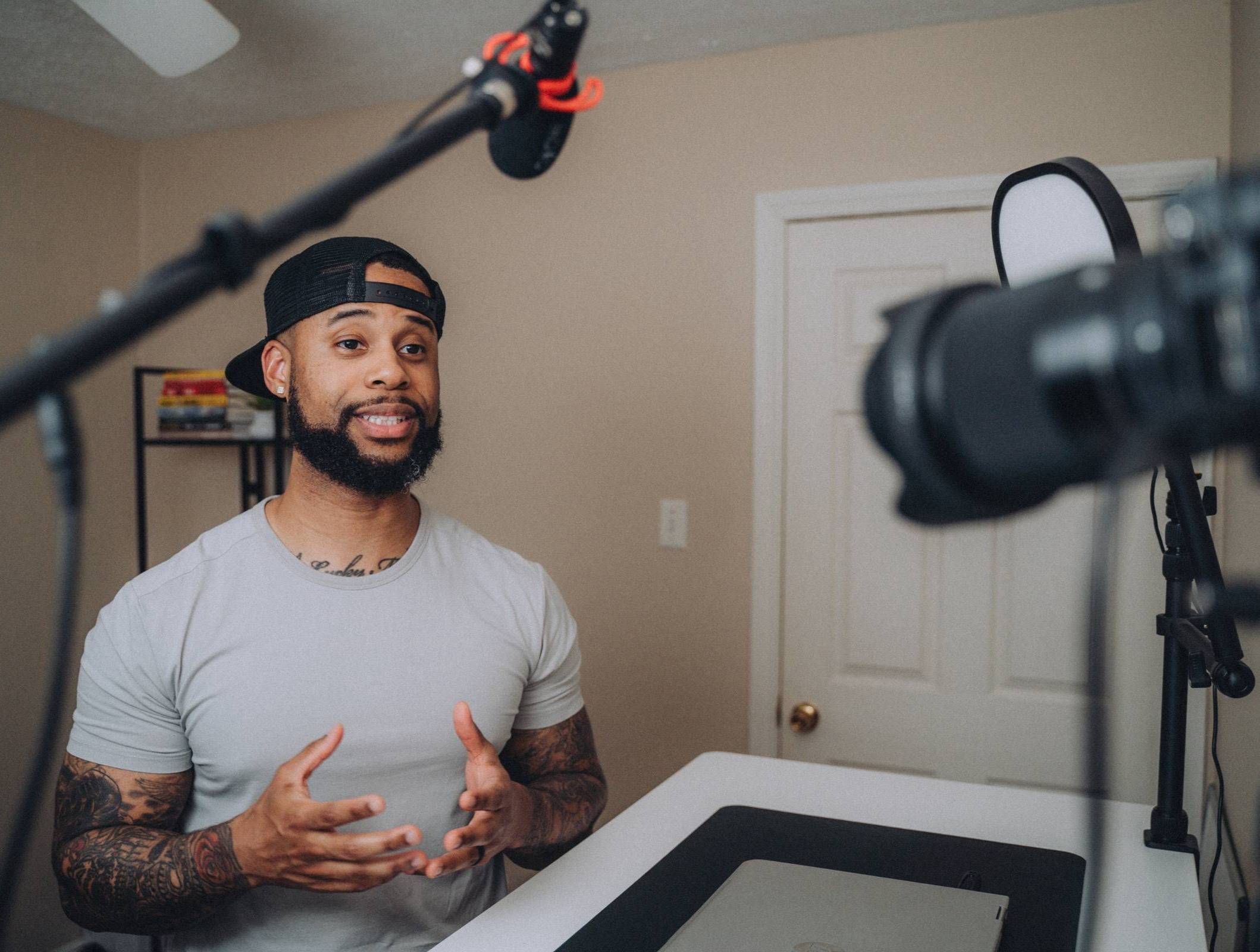 A man wearing a backward cap and a light gray t-shirt is speaking in front of a camera in a home studio. The room has beige walls, a closed door, and a shelf with books in the background. A microphone and camera equipment are visible in the foreground, indicating a video or podcast recording setup. He has tattooed arms and is gesturing with his hands while speaking.