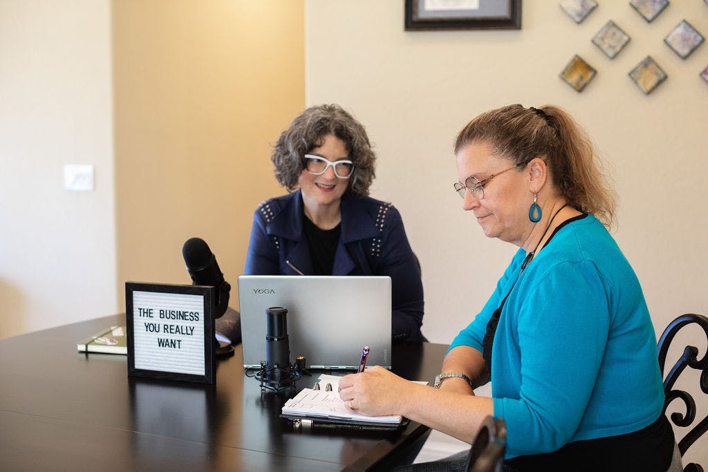 Tonya and Gwen outline their next podcast episode. Tonya sits in front of her laptop, while Gwen writes in her notebook.