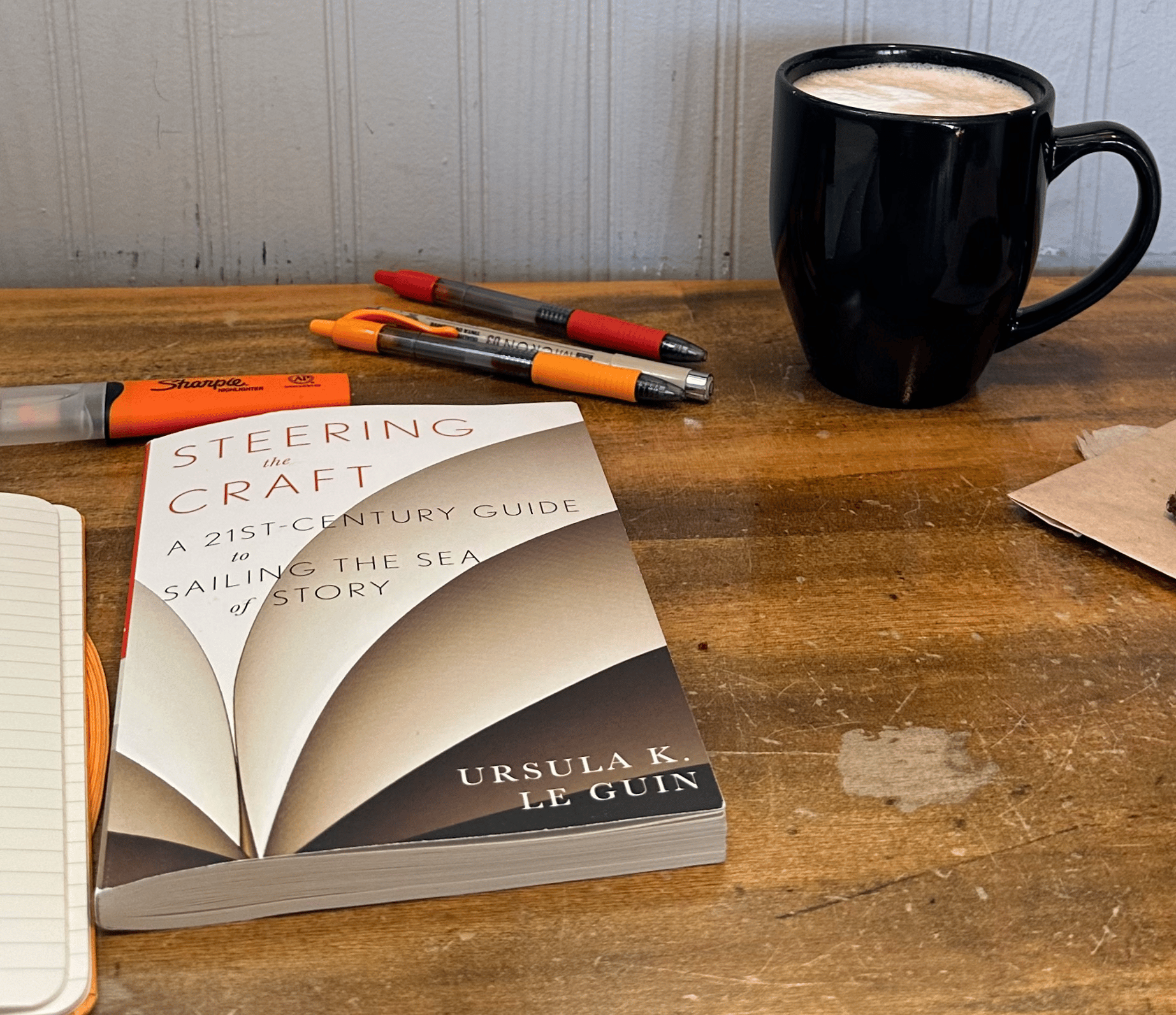 A book titled Steering the Craft by Ursula K. Le Guin on an antique desk, along with a mug of coffee, multicolored pens, and an orange highlighter.