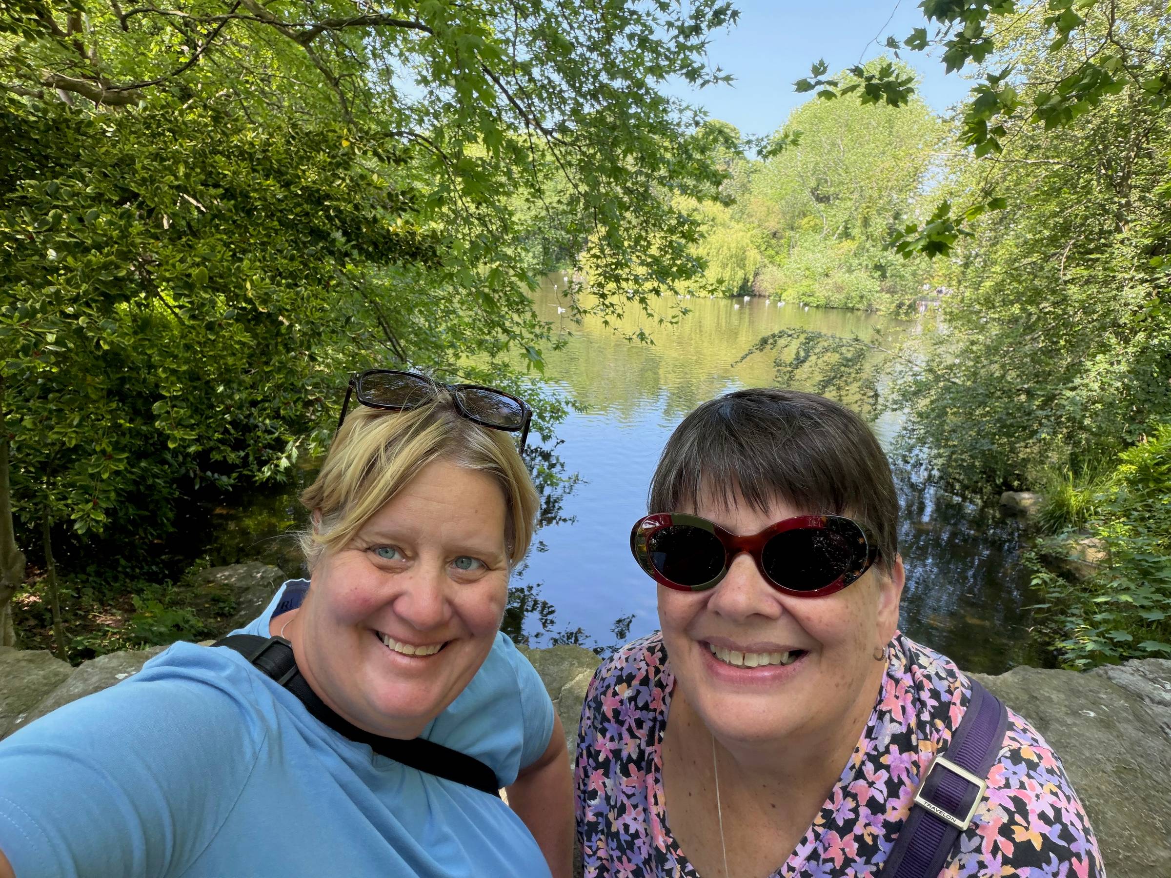 Two women smiling in a selfie at St. Stephen's Green, Dublin, with a sunlit lake and lush green trees reflected in the calm water behind them
