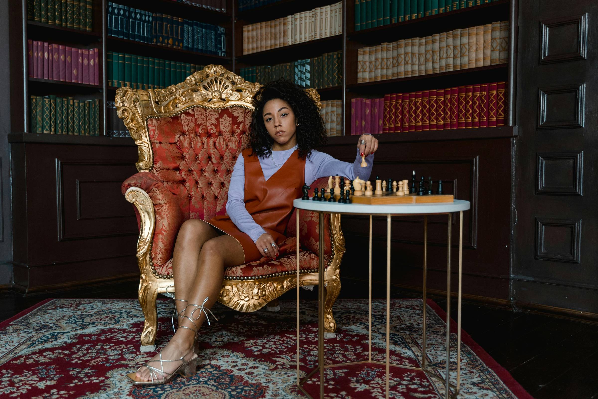 A woman sits confidently in an ornate gold armchair in a richly decorated library, holding a chess piece above a chessboard on a small table. Floor-to-ceiling bookshelves and a patterned rug frame the scene.