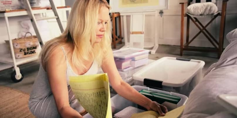 Pamela Anderson sits cross-legged on the floor, flipping through yellow legal pads from a storage bin, surrounded by clear plastic tubs filled with journals and notebooks. A quiet moment of sorting through a life once lived in the margins, echoing the ess