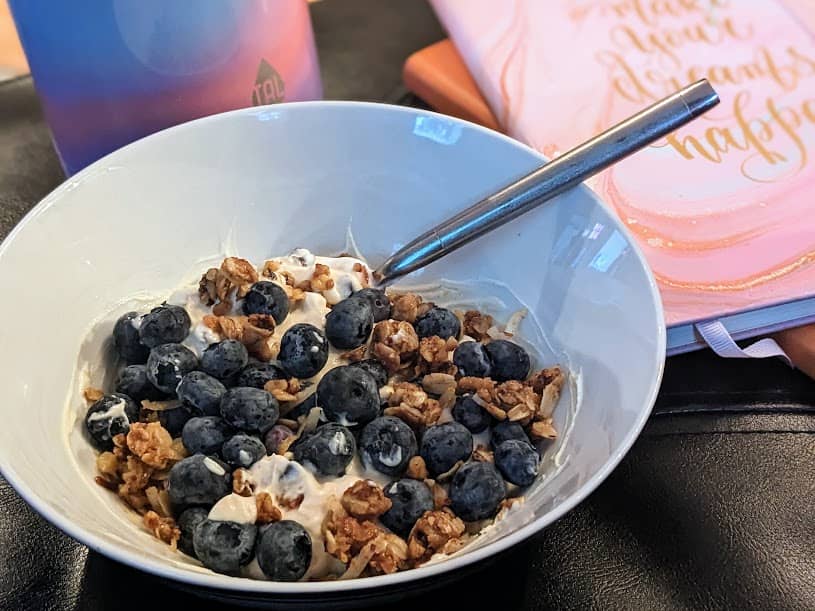 Bowl of Greek yogurt topped with granola clusters and fresh blueberries, a metal spoon resting in the bowl. In soft focus behind it are a pink orange gradient water bottle and spiral notebooks, one with gold script, on a dark table.