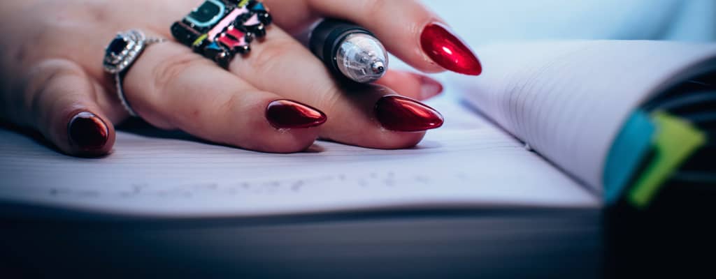A close-up photo of a hand with long, glossy red nails and ornate rings gripping a black pen. The hand rests on a white notebook with faint writing lines and some illegible cursive writing visible. The foreground is dramatically lit, with a blurred backgr