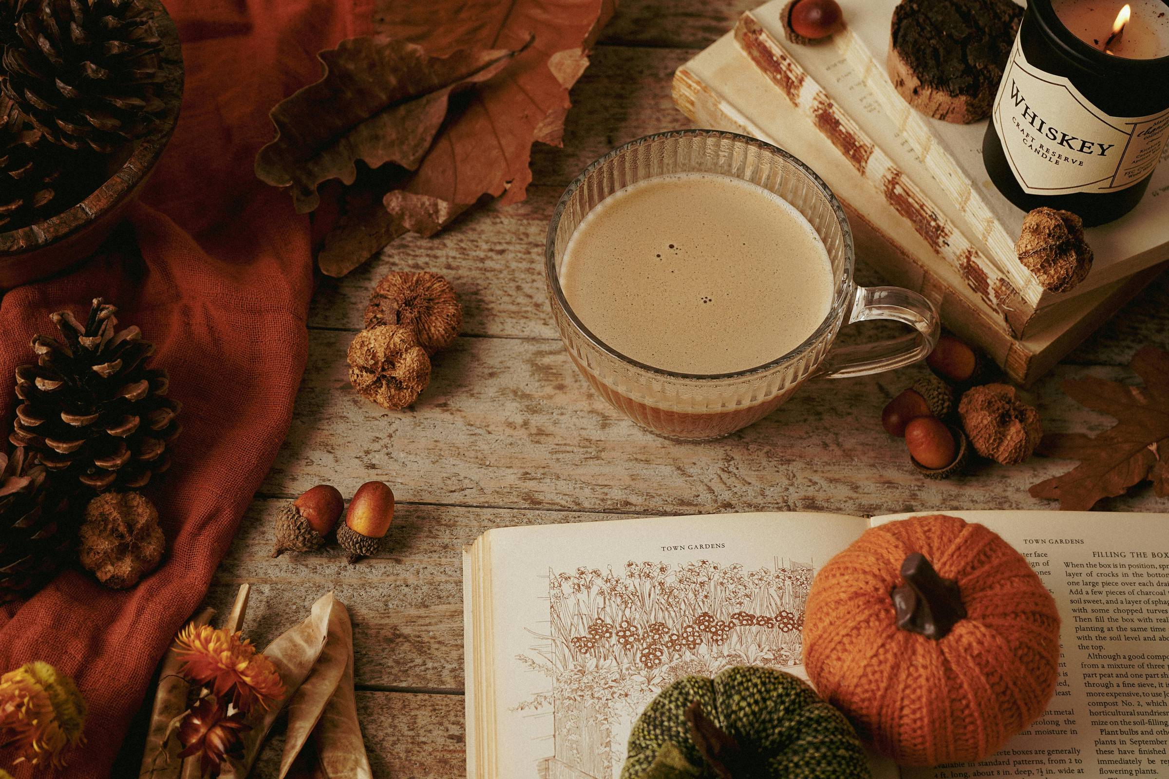 Warm fall still life on a wooden table: a glass mug of creamy coffee beside pinecones, acorns, dried leaves, an open vintage book, a candle labeled Whiskey, and two small knitted pumpkins. Cozy browns and oranges evoke pumpkin spice season.