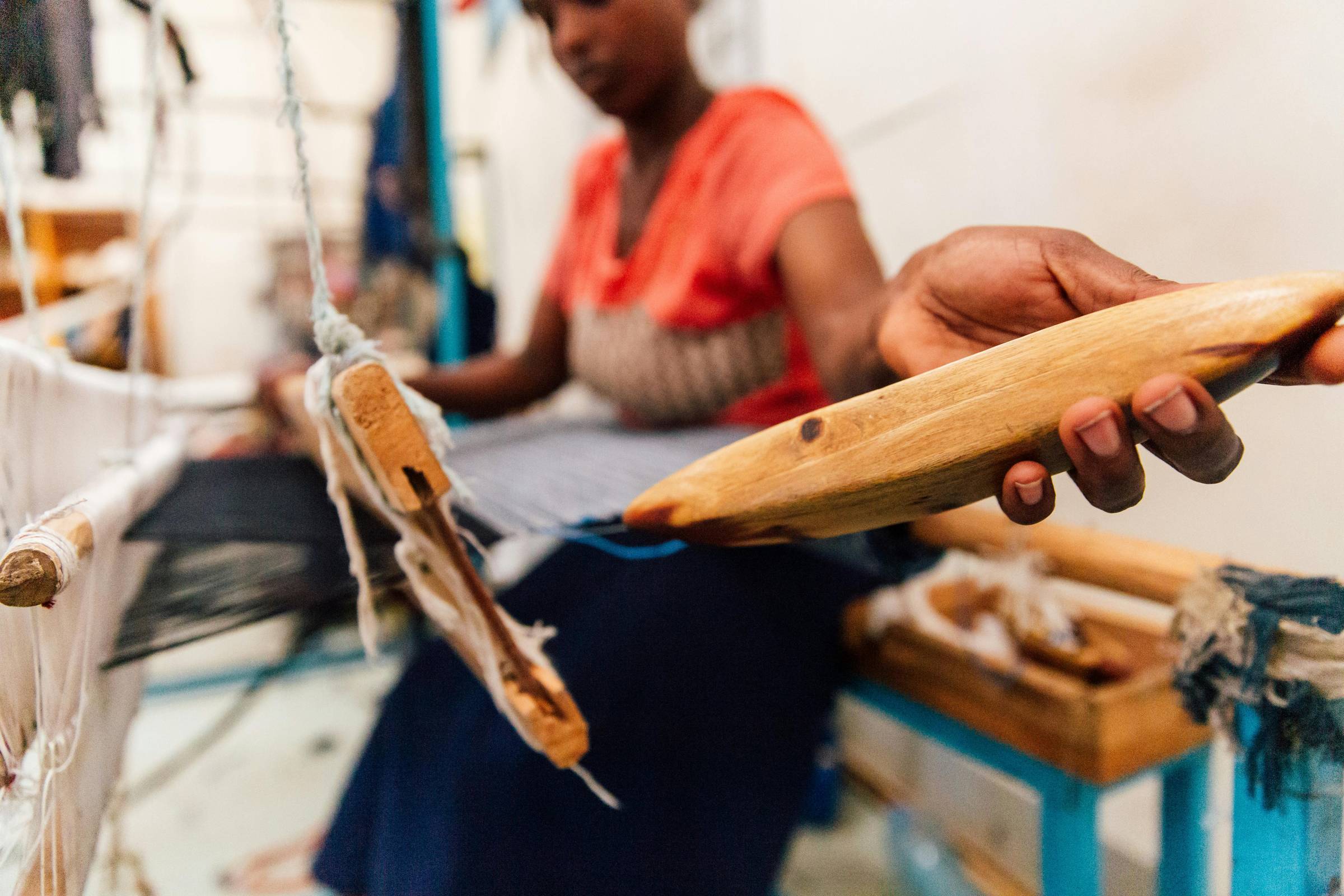 A close-up of a woman’s hand holding a wooden weaving shuttle mid-motion on a loom. Her face is out of focus in the background as she threads vibrant blue yarn through a half-woven textile. A powerful, grounded image of craft in action — every thread pull