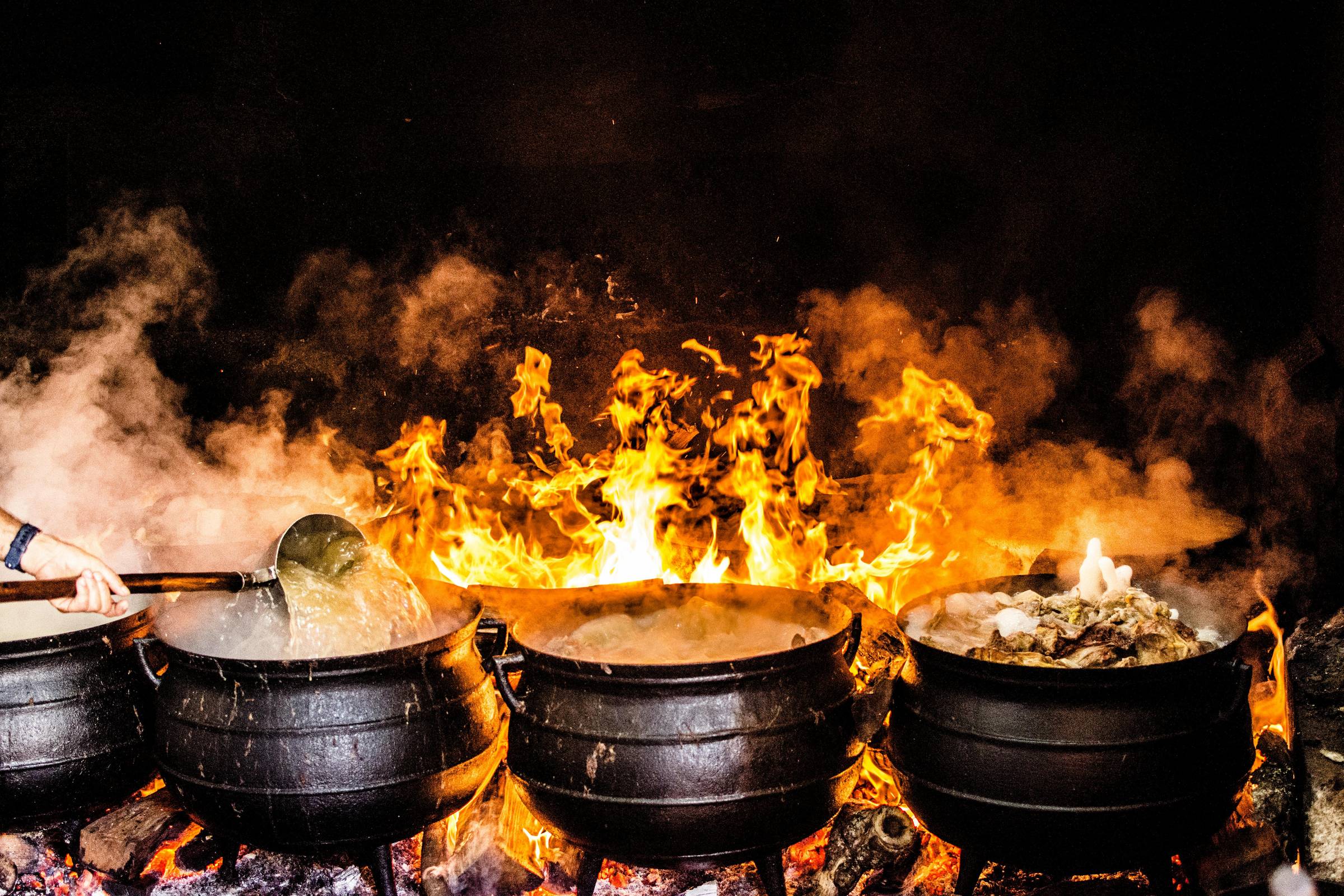 Four large black cauldrons boil over open flame, with steam rising and fire roaring beneath them. A hand stirs one pot with a long-handled ladle. The intense heat and chaos evoke both ancient kitchens and the creative process: unmeasured, elemental, and a