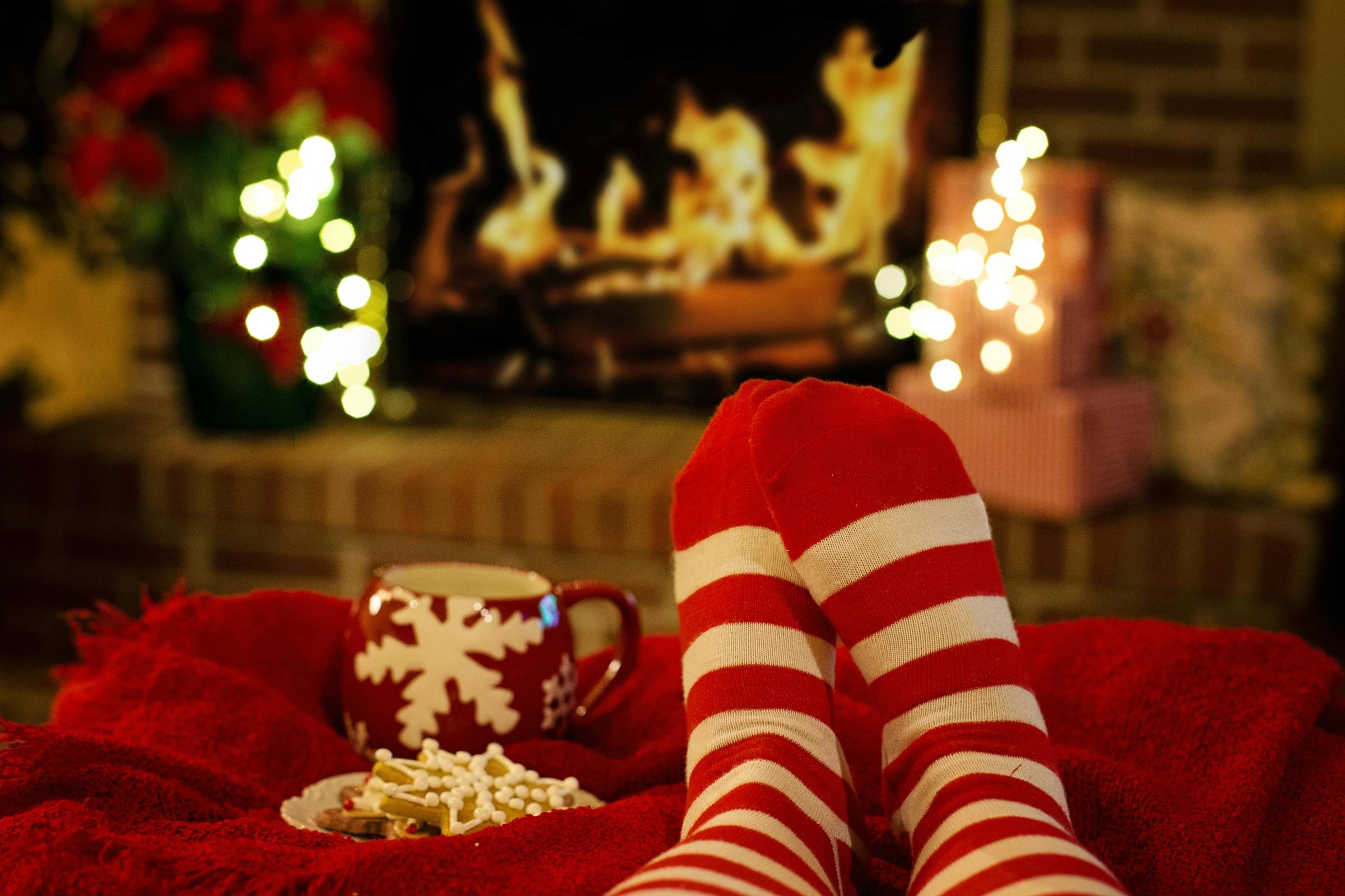 Person wearing red-and-white striped socks reclines near a lit fireplace, with a red mug, marshmallows, and soft holiday lights creating a cozy indoor winter scene.