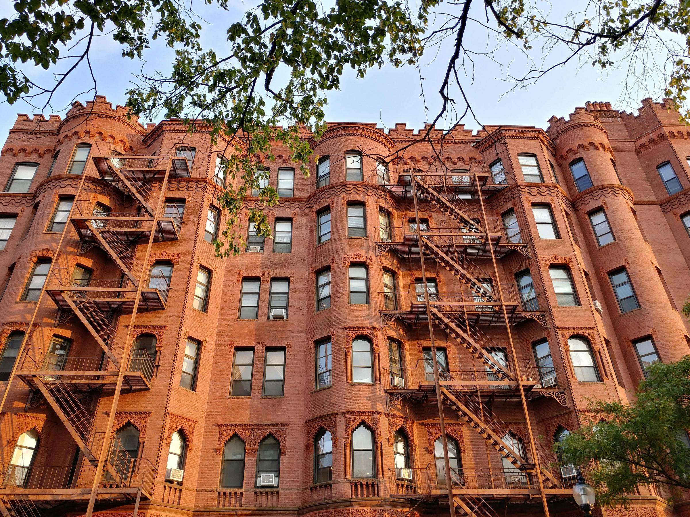 A red brick tenement-style building with elaborate ornamentation and two iron fire escapes zigzagging across its facade. The structure features arched windows, decorative brickwork, and castle-like towers, blending functionality with unexpected beauty. A 