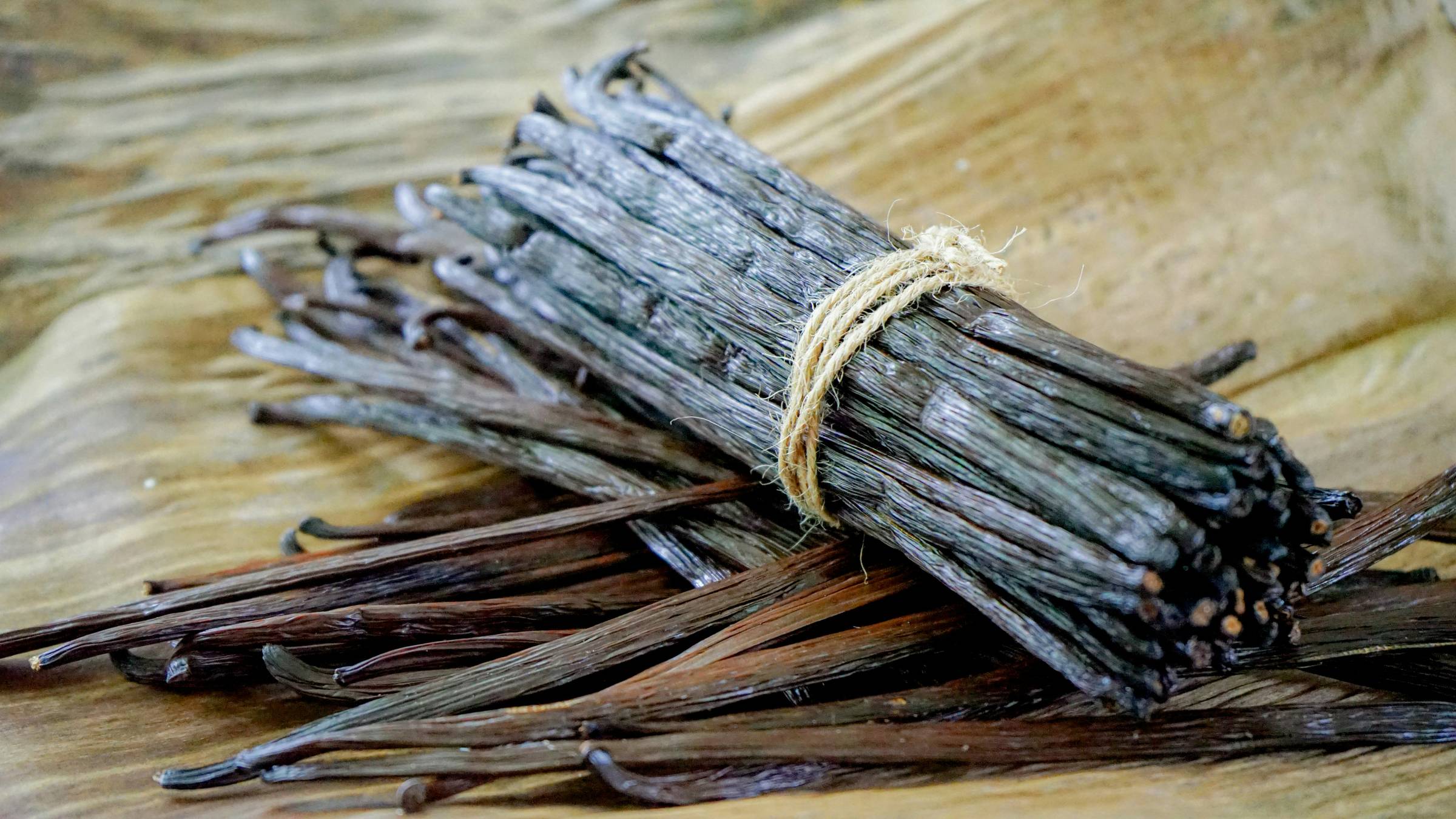 A bundle of dried vanilla beans tied with twine rests on a wooden surface. The dark, glossy pods are slightly wrinkled and textured, with some loose beans scattered around the bundle. Photographed in Sri Lanka.
