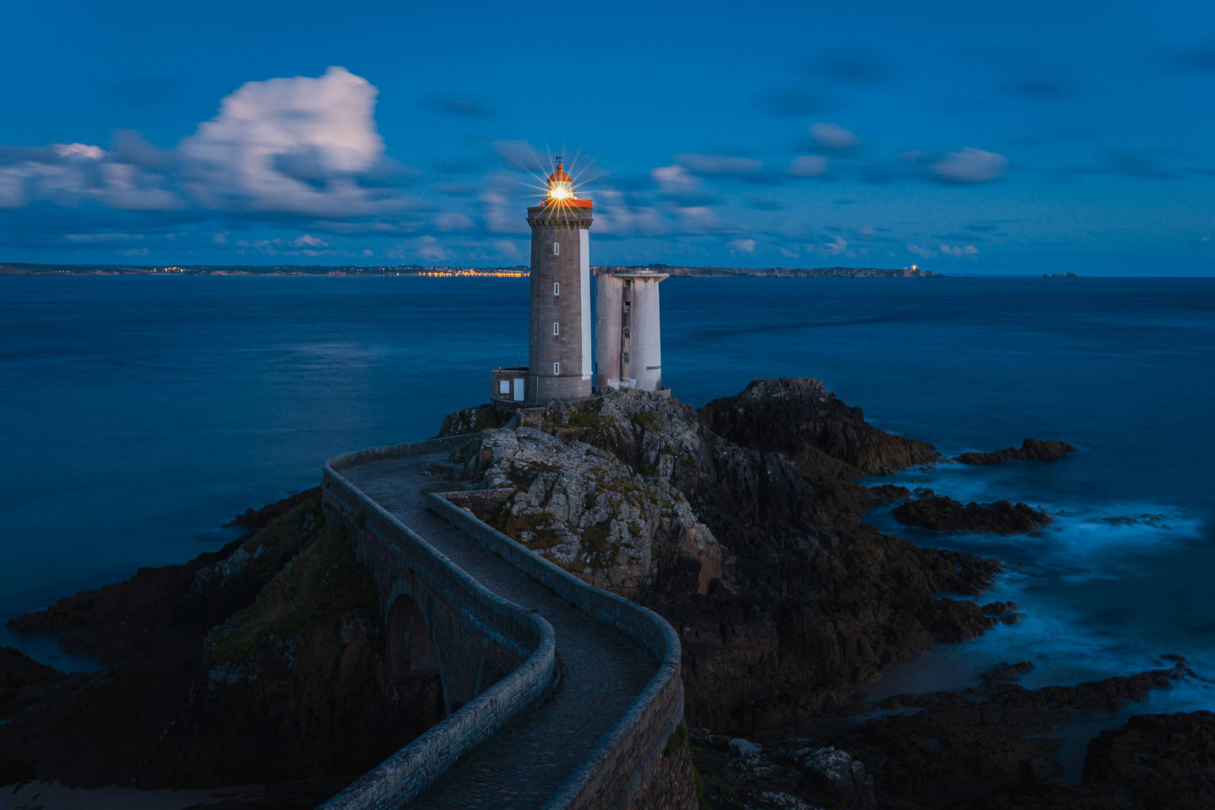 A stone lighthouse glows steadily at dusk on a rocky outcrop, connected to the mainland by a curved cobblestone path. The sea is calm, the sky soft with clouds, and the light cuts quietly through the blue,like a beacon meant not to shout, but to guide. 