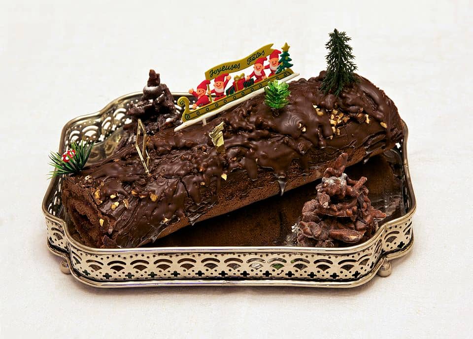 Chocolate Yule log cake coated in glossy chocolate icing, decorated with miniature trees, pinecones, and a small festive figurine scene, served on an ornate metal tray.