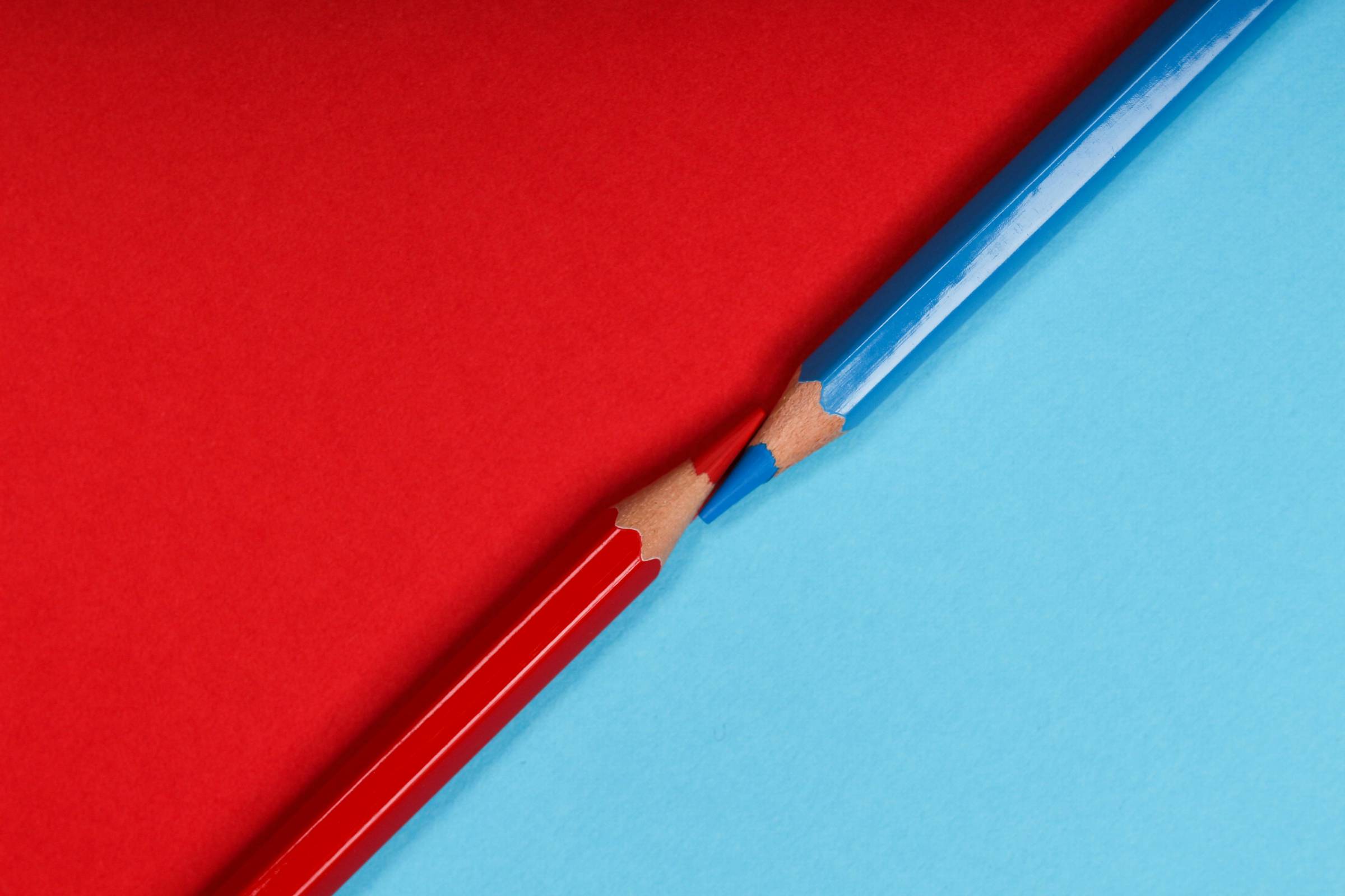A close-up image of two sharpened colored pencils—one red, one blue—pointing at each other diagonally. The red pencil lies on red paper, and the blue pencil lies on blue paper, forming a crisp contrast at their intersection.
