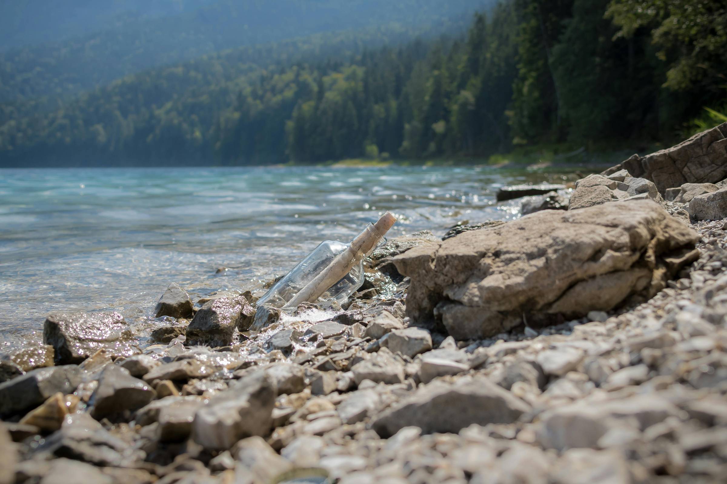 A glass bottle with a cork and rolled-up paper inside lies among wet rocks at the edge of a lake. The shoreline is rocky, with forested hills and misty mountains in the background.