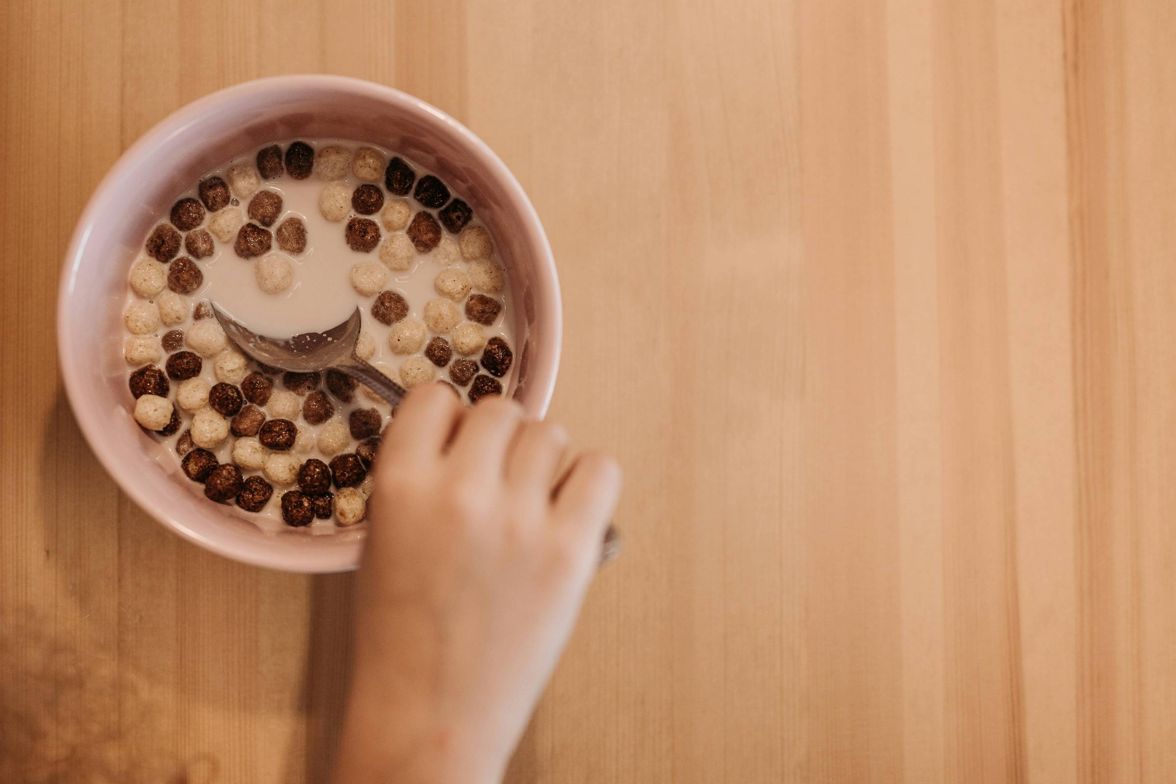 A hand holding a spoon over a bowl of cereal with chocolate and peanut butter puffs floating in milk, on a light wooden table.