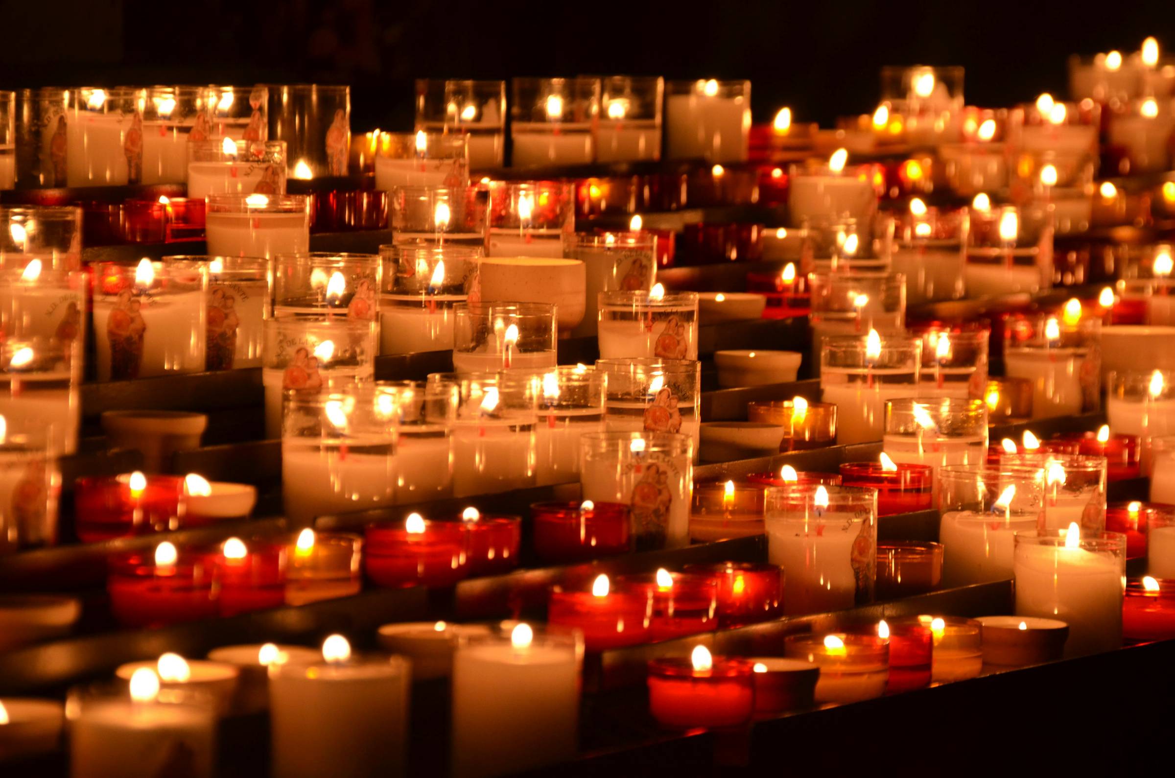 Dozens of small red and white votive candles in glass holders burning in rows, their soft flames creating a warm, flickering glow in the dark