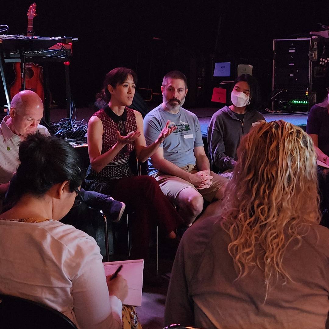 Vienna sits in a chair in front of a stage, keyboard and guitar visible in the background, in a circle of people listening or taking notes. She is gesturing with both hands held at chest level, palms up.