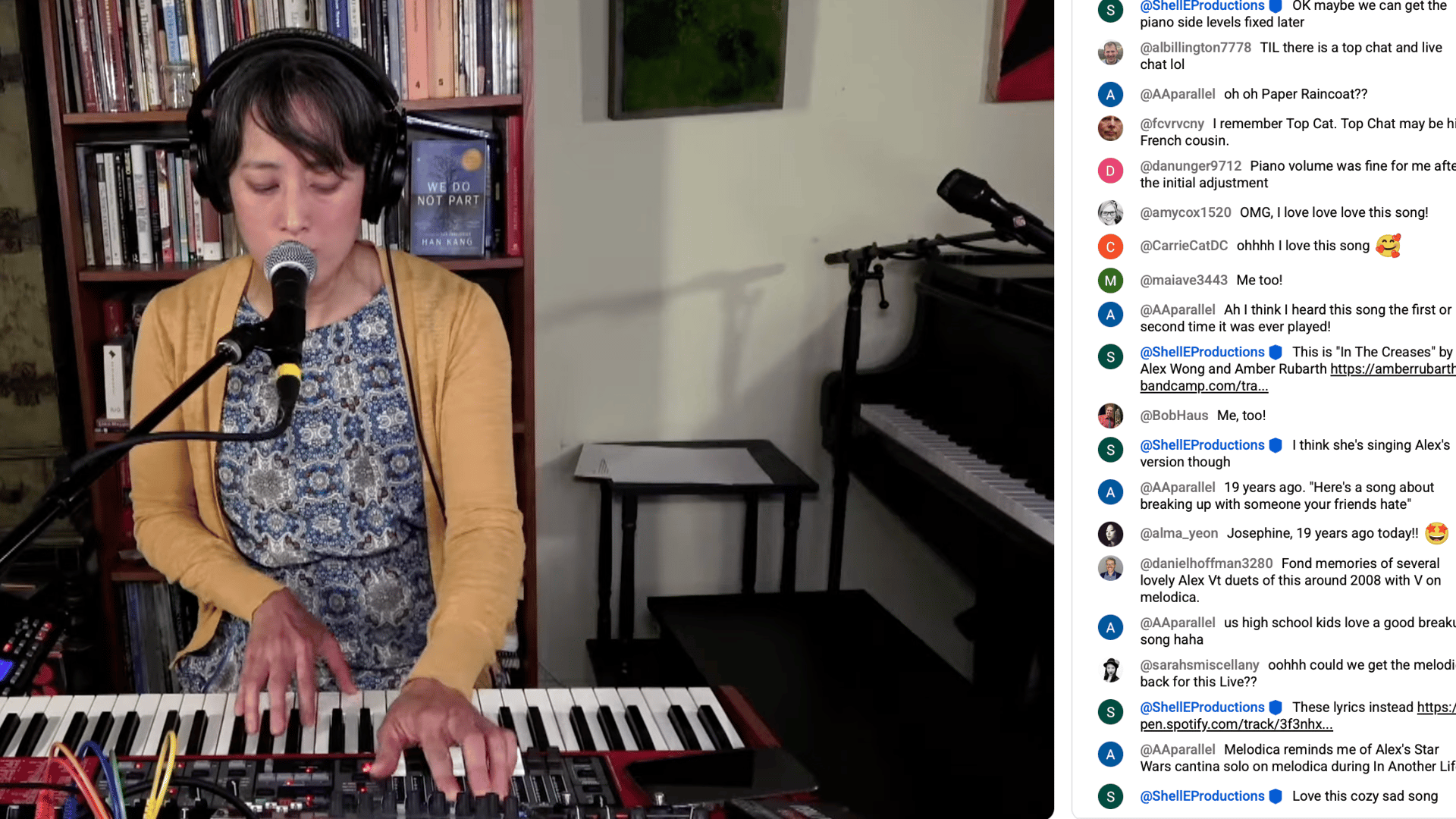 Screenshot of Vienna performing in a livestream, singing into a microphone and playing a red keyboard while pressing other buttons with her left hand. She wears headphones, a yellow cardigan and a blue-and-white patterned dress. The live chat is visible t