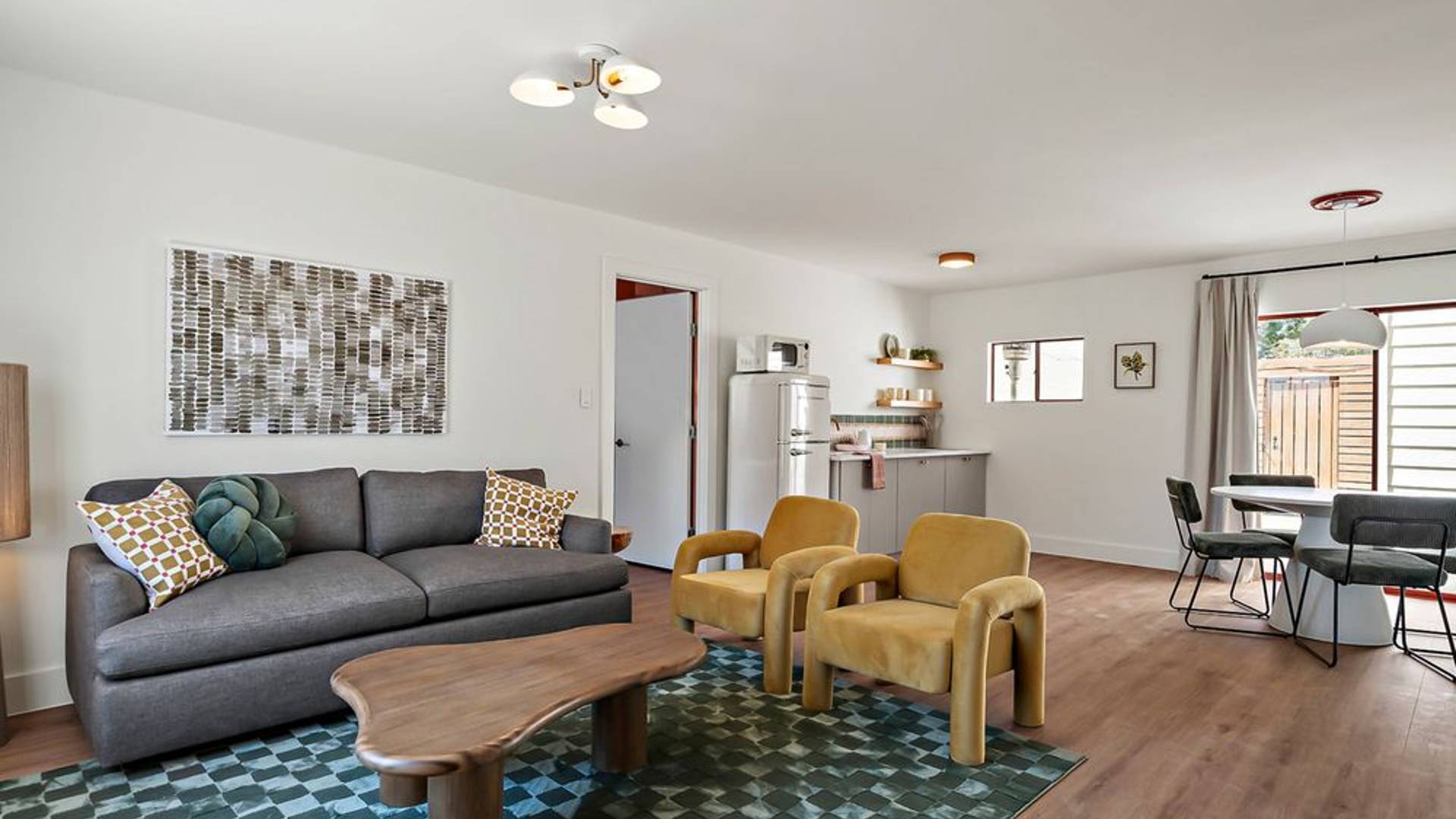Promotional photo of Hacienda Del Rio, showing the living room and kitchenette of a suite. The decor and color palette is modern and relaxed: a gunmetal gray couch, curved wooden coffee table, mustard yellow armchairs, white walls and natural wood floors with checkered teal rug. A patio is visible through the sliding glass doors on the right, and the room is full of natural light.
