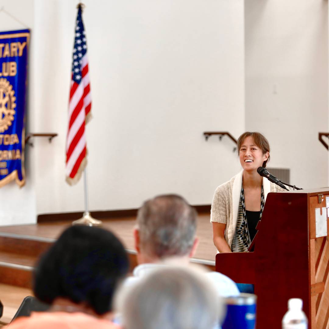 Vienna sits at an upright piano, smiling, with audience members' heads visible in the foreground, and a Rotary Club and American flag visible in the background.