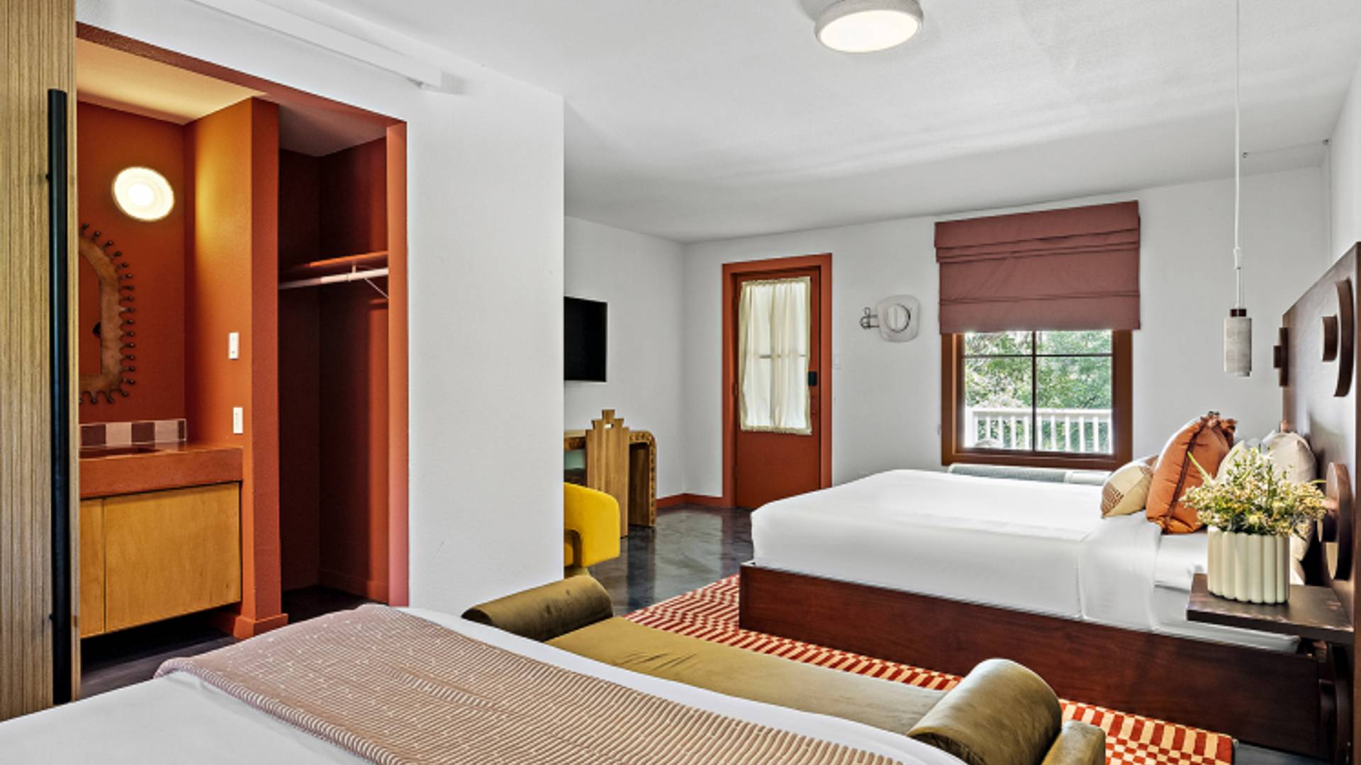 Promotional photo of Hacienda Del Rio, showing a king + queen bed double room. The color palette and decor is modern and warm: deep orange, dark wood, white walls and sheets, muted yellow and green accents.