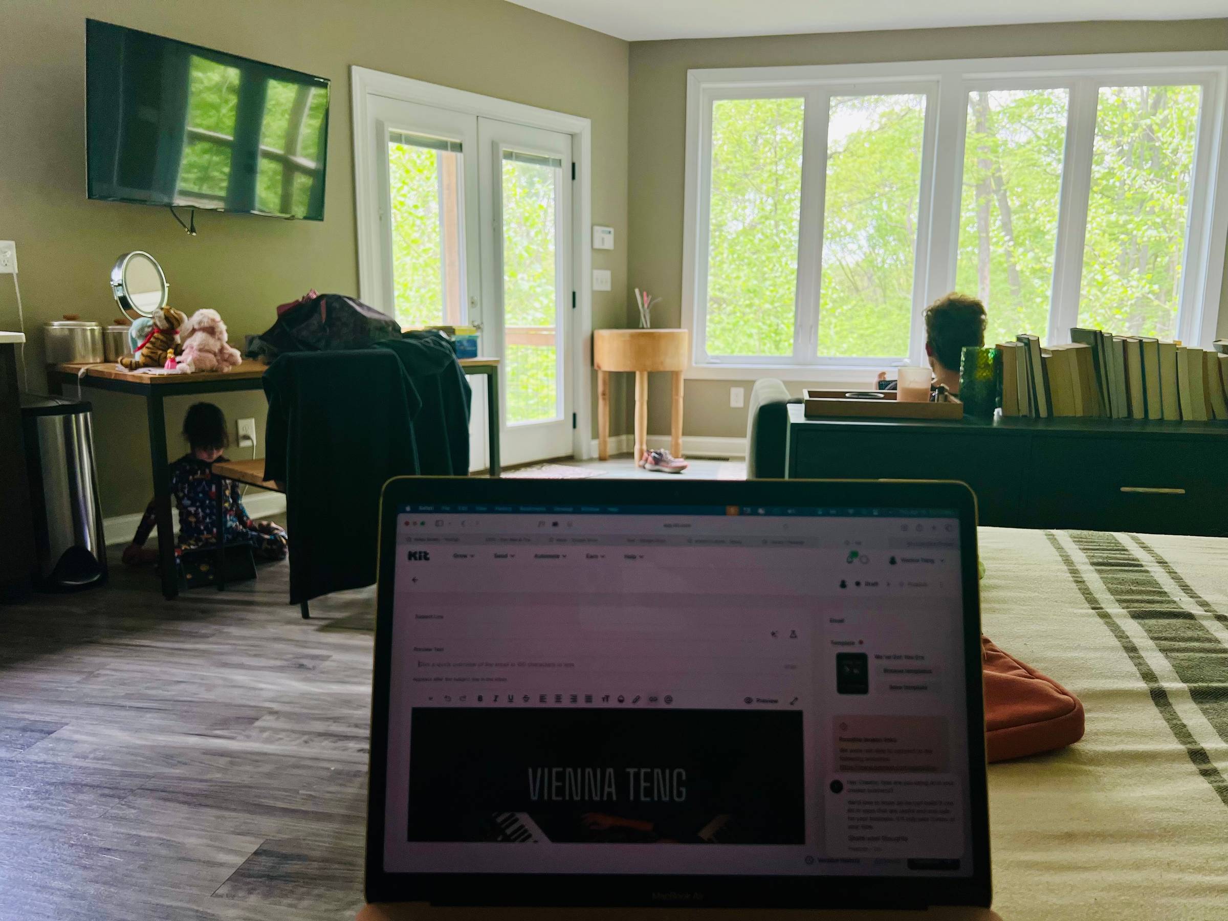 A laptop screen is in the foreground, showing a newsletter being composed, while the writer (presumably Vienna) sits on a bed facing windows filled with tree foliage. The back of a man's head, presumably Jacob, is visible on a couch facing the window as w