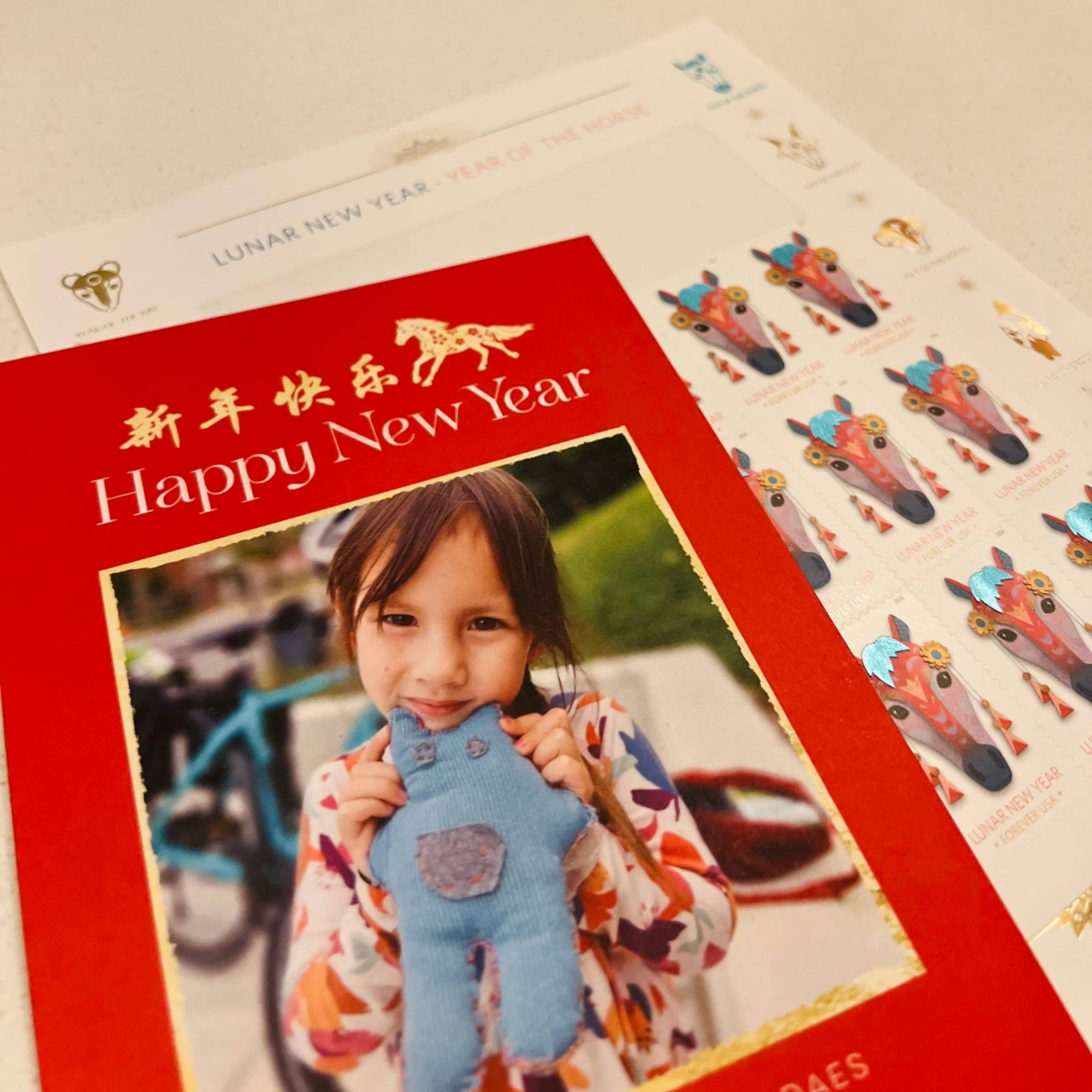 Photo of Vienna Teng's family's 2026 Lunar New Year Card. It features a photo of her daughter Arcadia holding a roughly hand-sewn doll that she likely made herself. The Chinese characters for "happy new year" and an icon of a horse appear in yellow above the words "Happy New Year" in English, on a red background. A sheet of Year of the Horse Lunar New Year stamps is tucked behind the card.