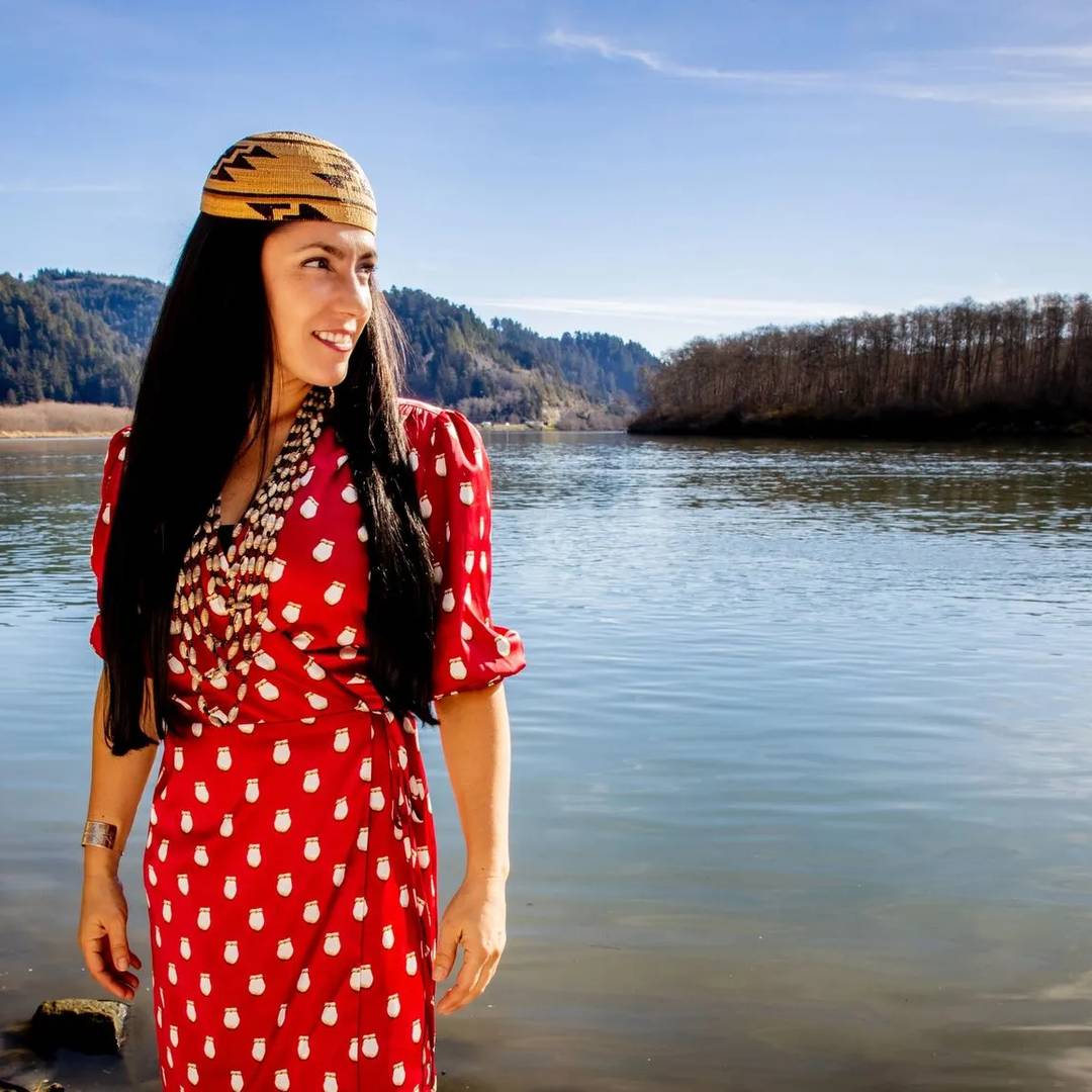 Amy Bowers Cordalis stands in front of the Klamath River, looking to the right, wearing her Yurok basket cap and beads and a bright red and white dress.