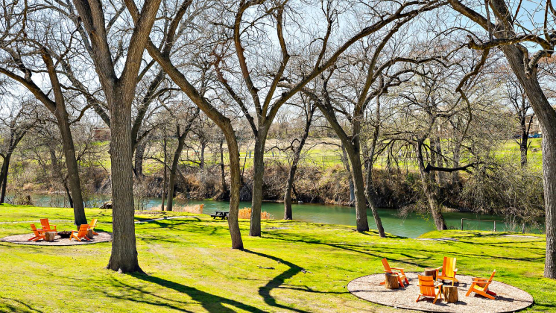 Promotional photo of Hacienda Del Rio, showing the grassy wooded area leading down to the Guadalupe River, including two sandy fire pit areas with orange Adirondack chairs.