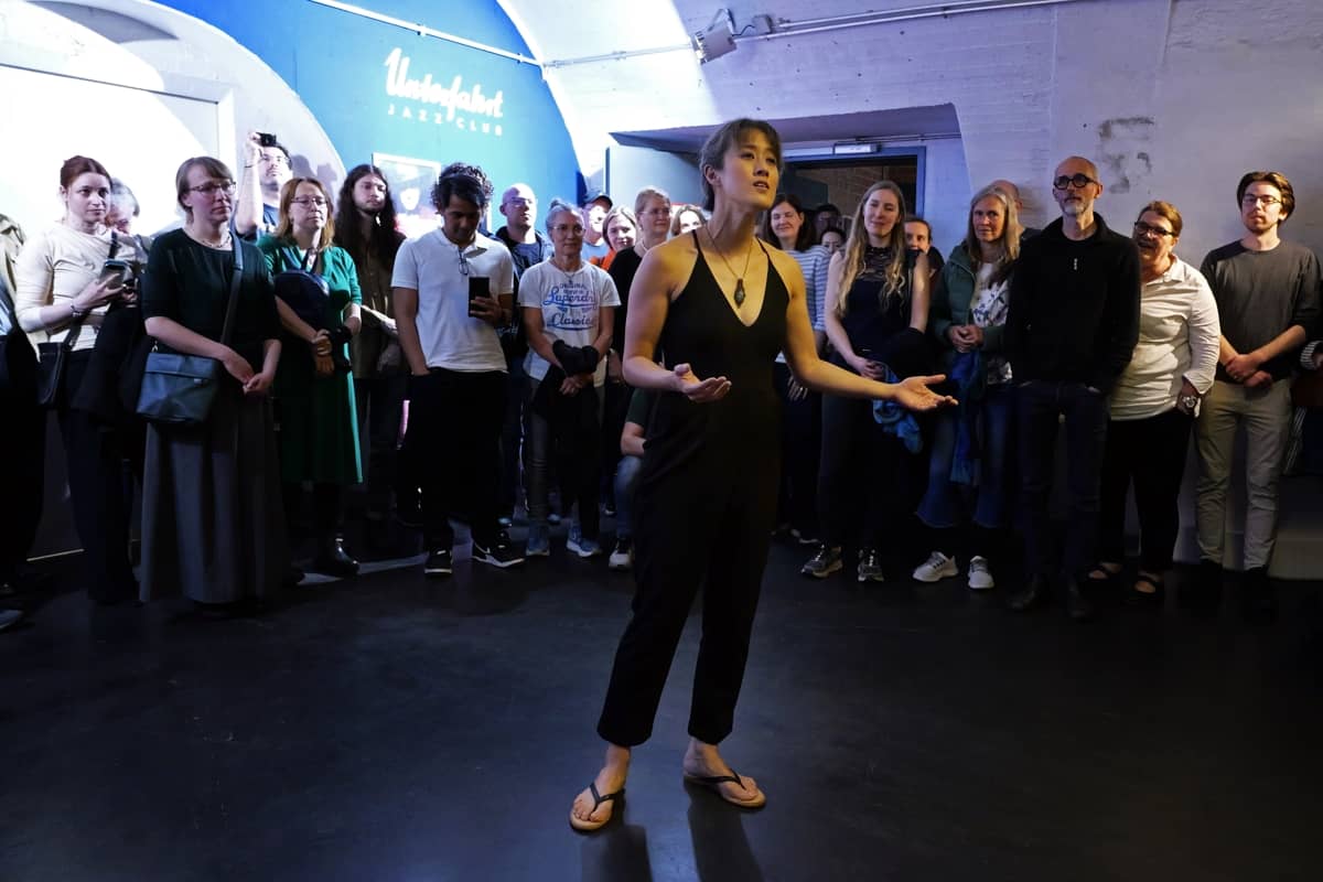 Vienna stands with hands outstretched in the middle of a circle of people listening (half the circle is shown), singing. She wears a black sleeveless jumpsuit and sandals. On a wall behind her, the words "Unterfahrt Jazz Club" are visible. 