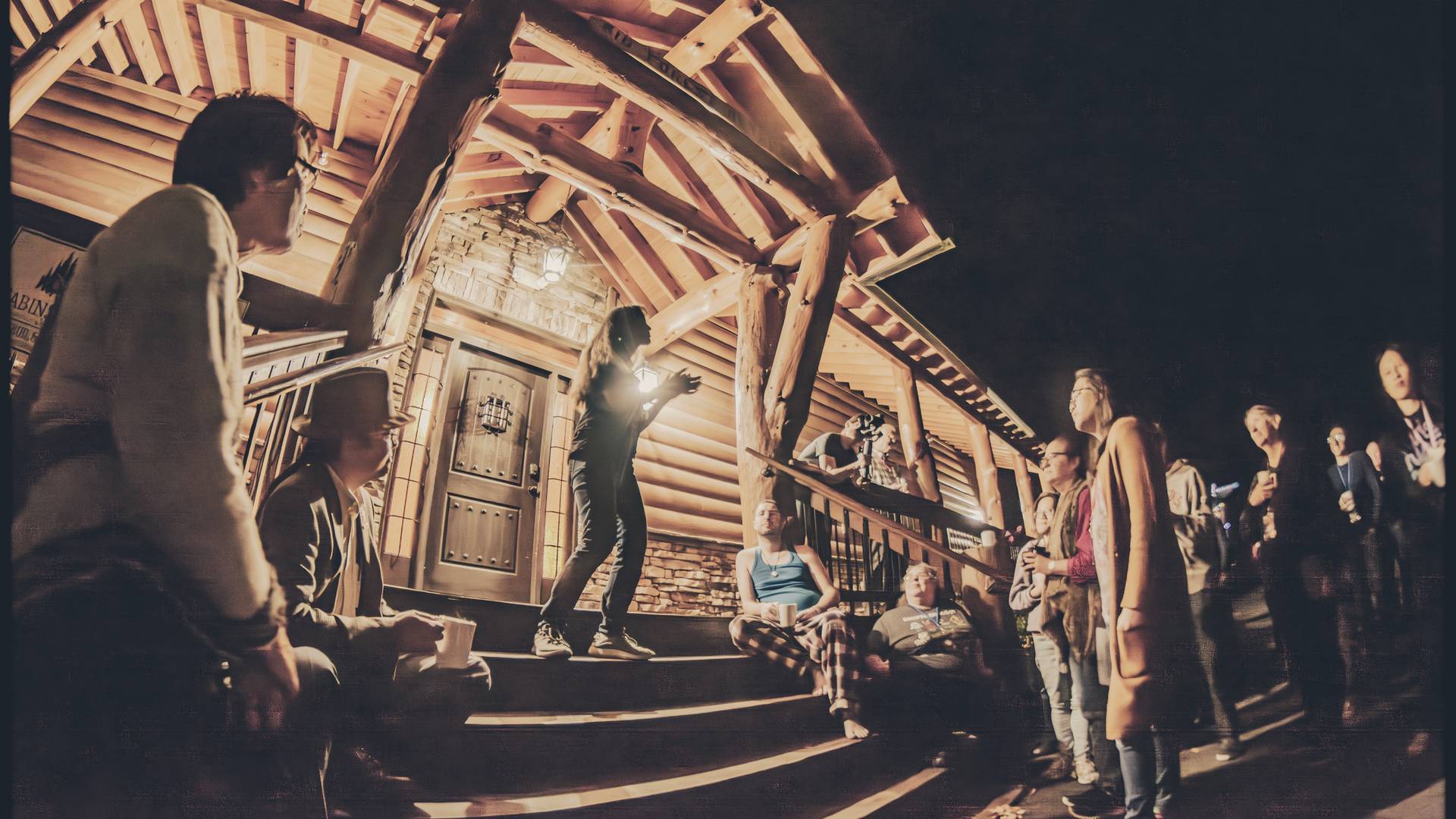 Photo from a previous Multitudes event, showing a nighttime event where Vienna is standing at the top of a set of porch steps, people gathered around her, singing and clapping or gesturing dramatically while speaking. 