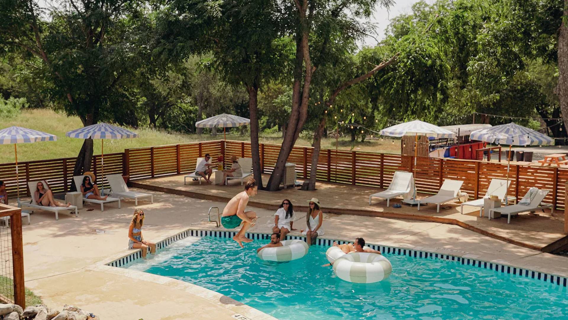 Promotional photo of Hacienda Del Rio, showing the pool area: an expansive deck with pool lounge chairs and umbrellas, shade from tall trees, and large white-and-gray inflatables in the pool. People are lounging beside and in the pool, with one person in midair, legs tucked for a cannonball jump into the water.