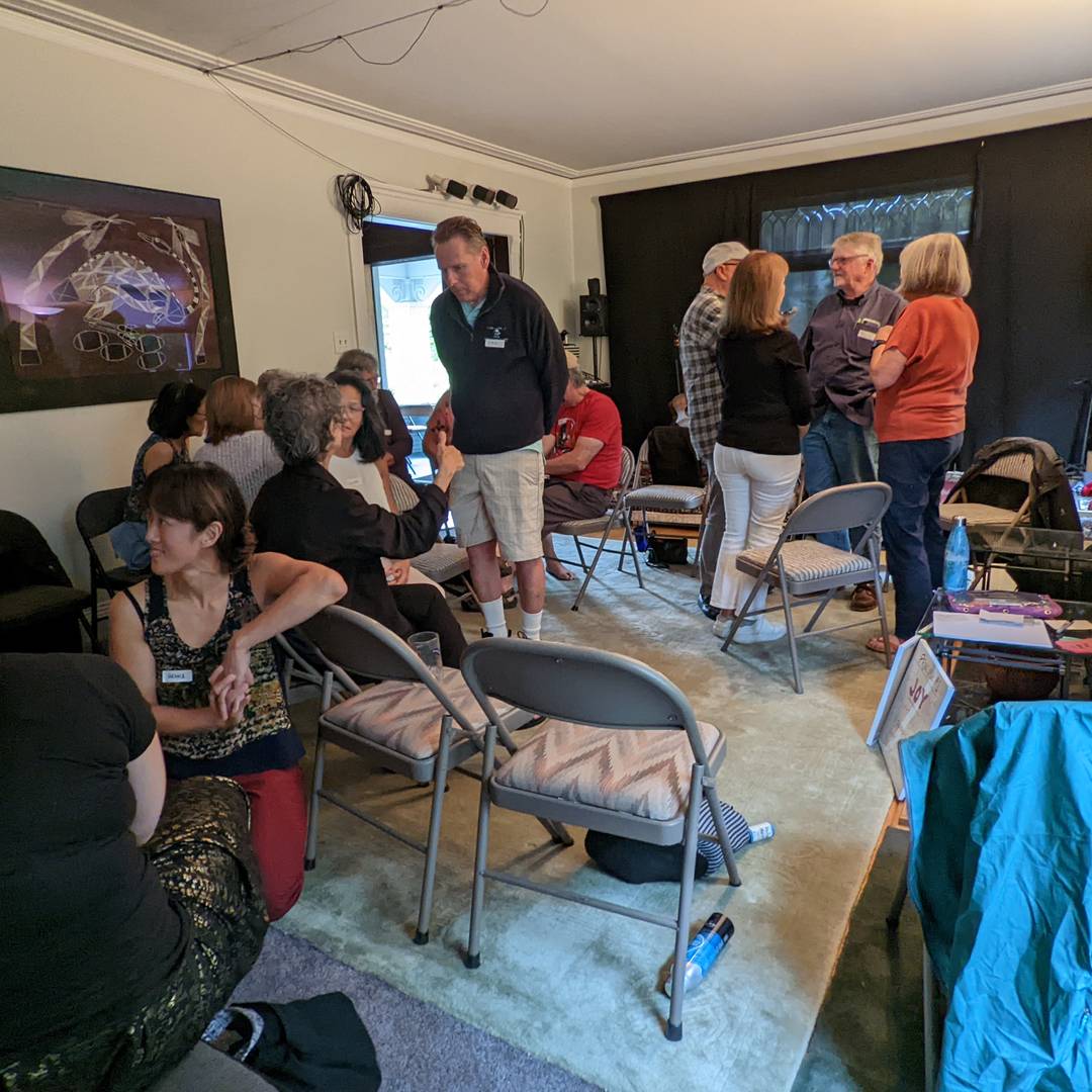 A roomful of people converse in small groups, sitting in or standing among folding chairs. Some people's name tags are visible on their chests, including Vienna, who crouches on the floor to participate in one group's discussion.