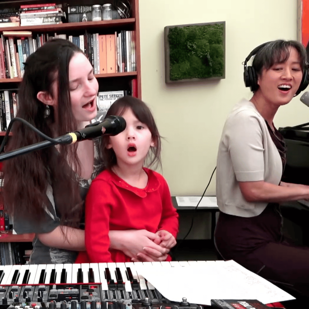 still from a livestream video. Vienna sits facing right playing a piano, wearing headphones and a short sleeve beige shrug sweater. Two other singers, her daughter and stepdaughter, sing into the microphone at her electronic keyboard station, facing the camera. Her daughter wears a bright red long sleeve top with a Peter Pan collar. Her stepdaughter wears long earrings and a gray-and-white flutter sleeve top, her hair tied back except for a section around her face. A bookshelf and a moss wall hanging are visible in the background.