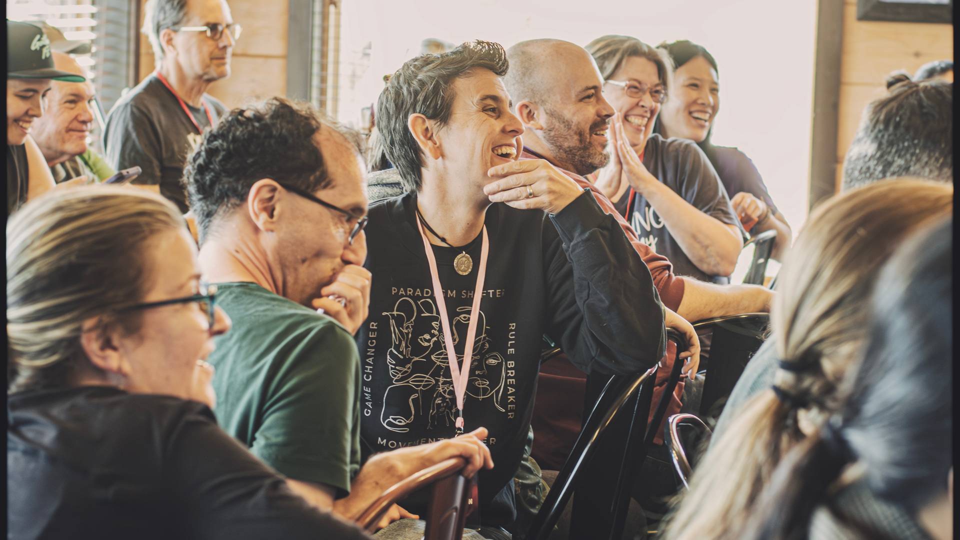 Photo from a previous Multitudes event, showing people seated closely together and looking to the right, laughing and smiling.