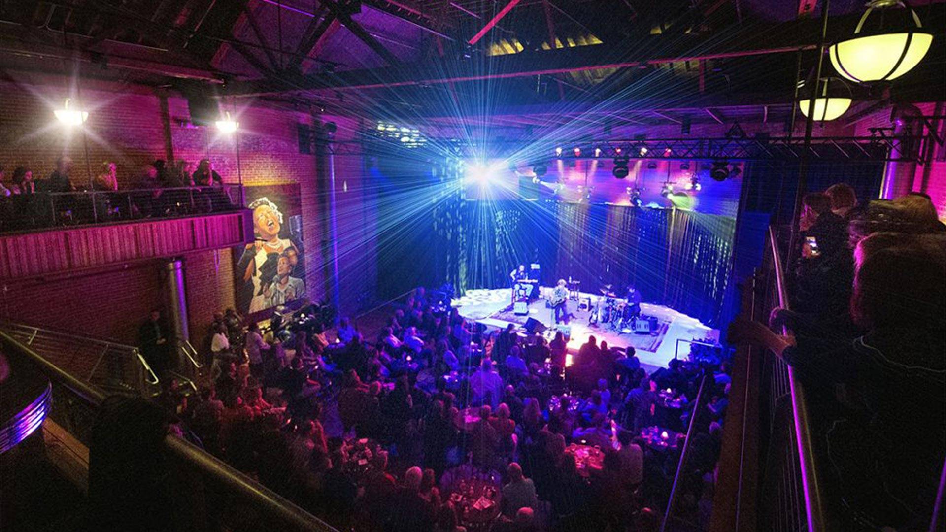 Photo taken from the right balcony of The Armory in Fort Collins. A mirrorball-like burst of light emanates from the stage, and the room is lit in magenta and purple hues.