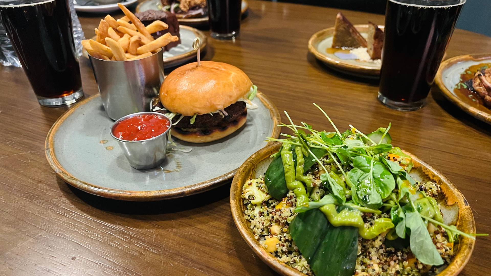 Photo of two plates of food on a wood grain table: a burger and fries with ketchup, and a grain salad topped with greens and a green dressing. Pint glasses of a dark beverage and other plates of food are also visible.