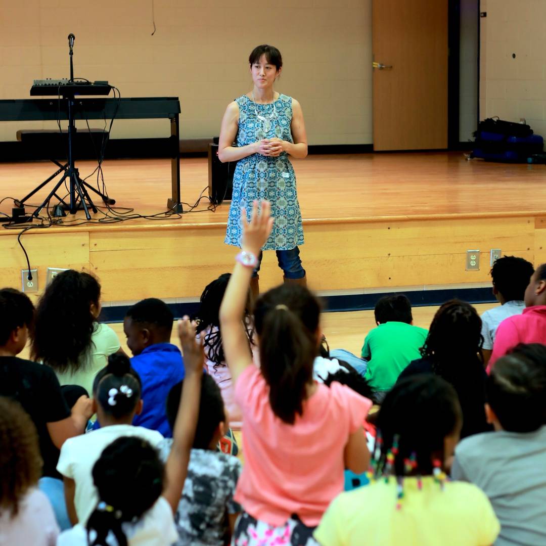Vienna stands on the floor in front of a stage, a keyboard visible behind her, looking at an audience of elementary-aged children, two of whom have their hands raised.