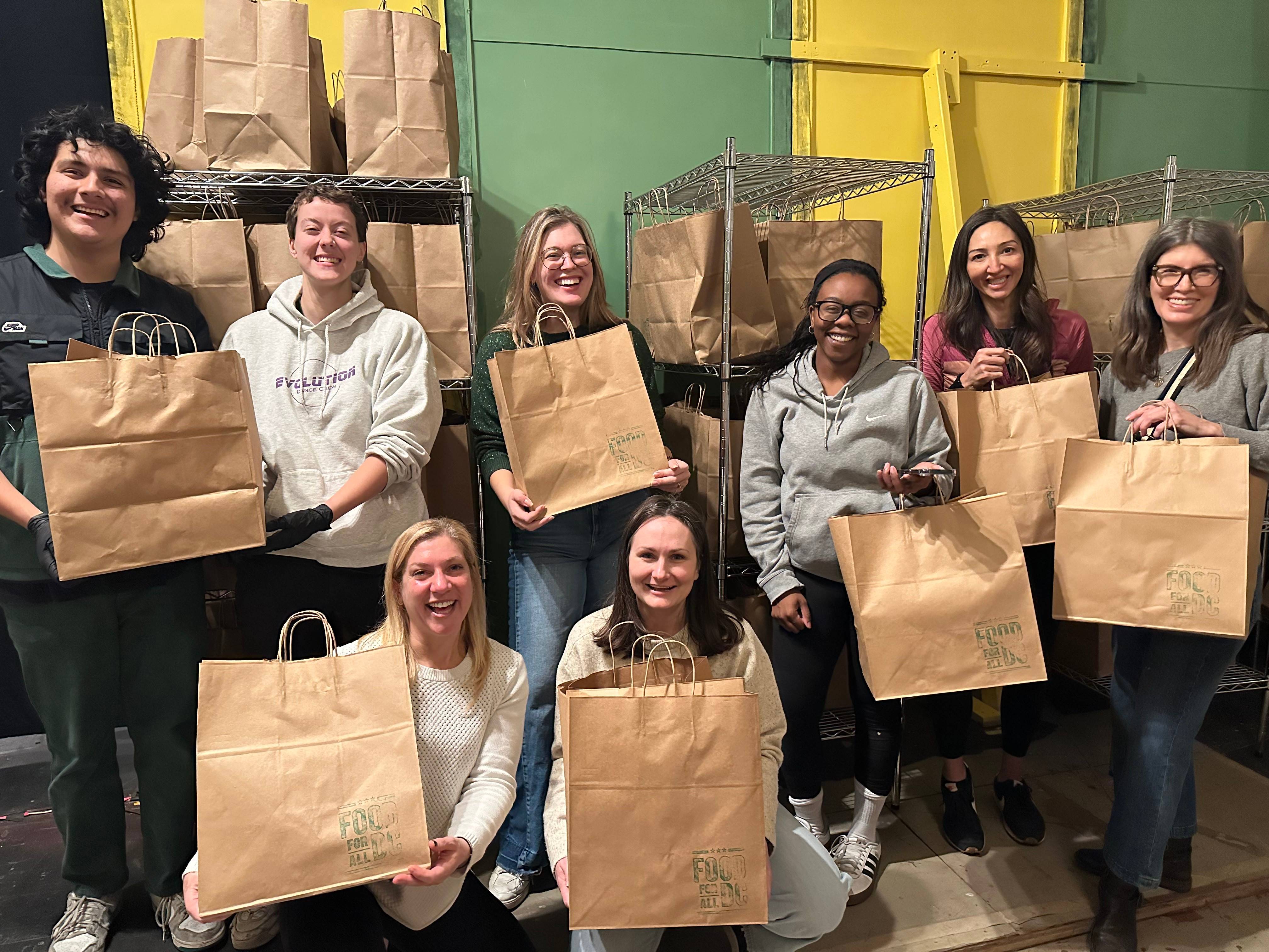 The image shows a group of eight people smiling and holding paper bags labeled "Food For All" in front of shelves stocked with similar bags. The setting appears to be a community food distribution or volunteer event, with a brightly colored background fea