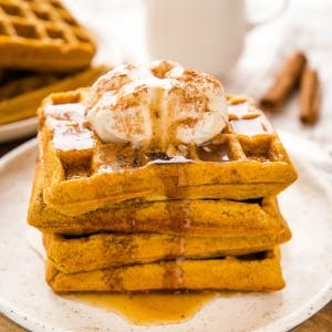 stack of pumpkin waffles, drizzled with syrup and topped with whipped cream