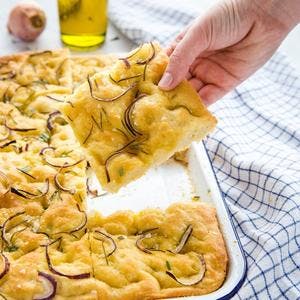 square slice of focaccia bread topped with roasted onions and rosemary being removed from a pan